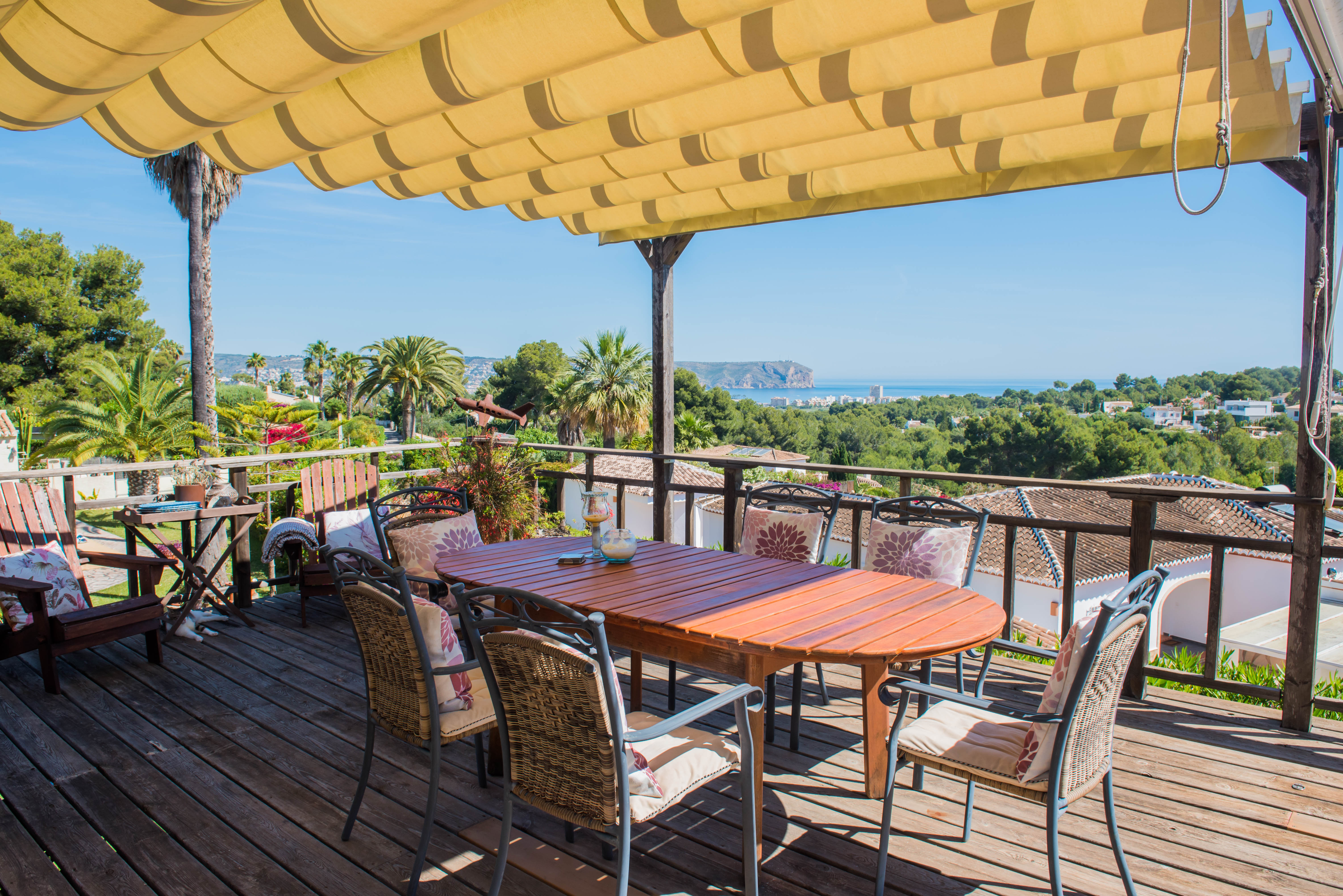 Holzterrasse mit Markise und Esstisch, mit Blick auf das Meer und üppiges Grün.