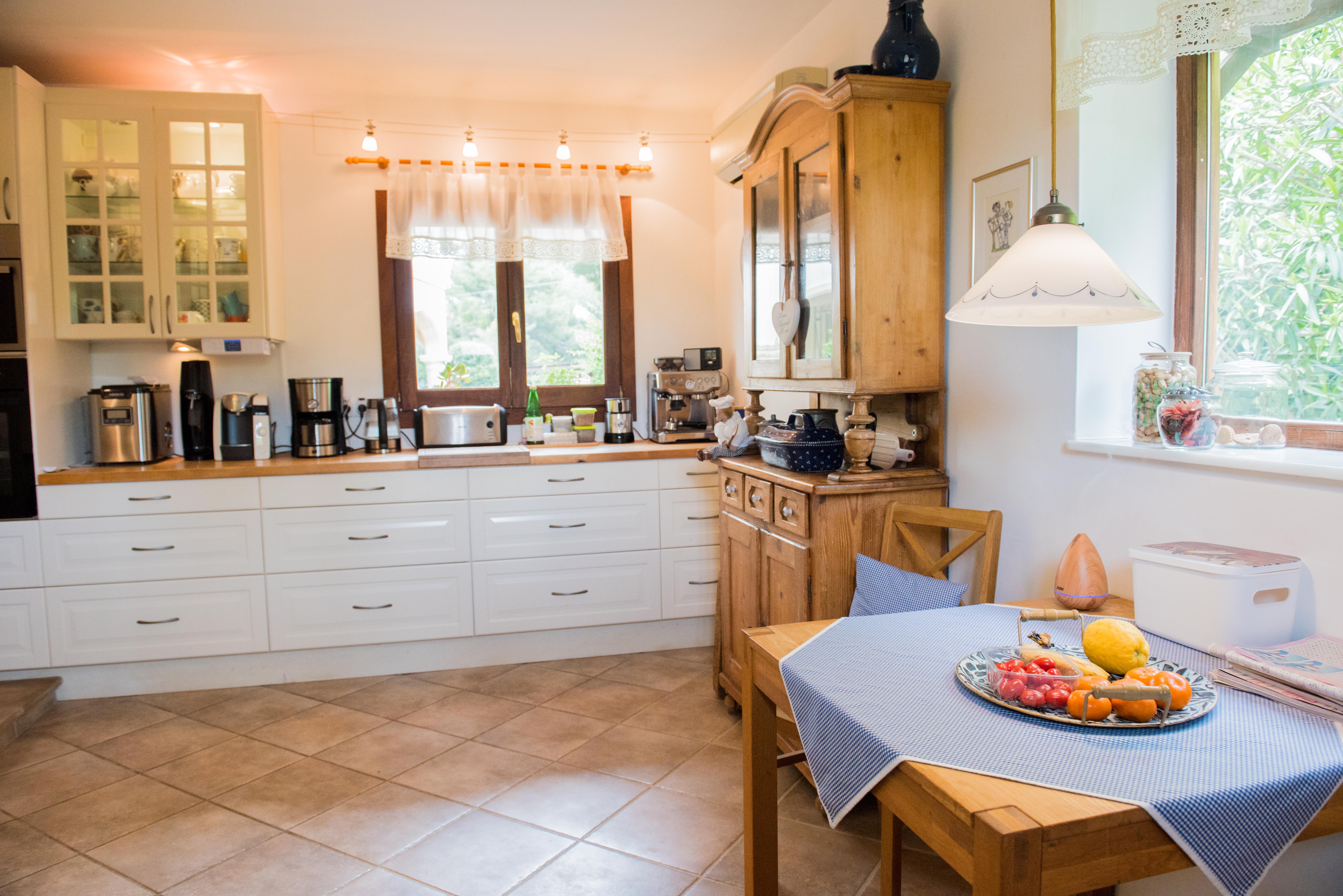 Rustikale Küche mit weißen Schränken, Holzarbeitsplatten und Essbereich. Mit klassischem Holzbuffet, Keramikfliesenboden und natürlichem Licht aus zwei Fenstern.