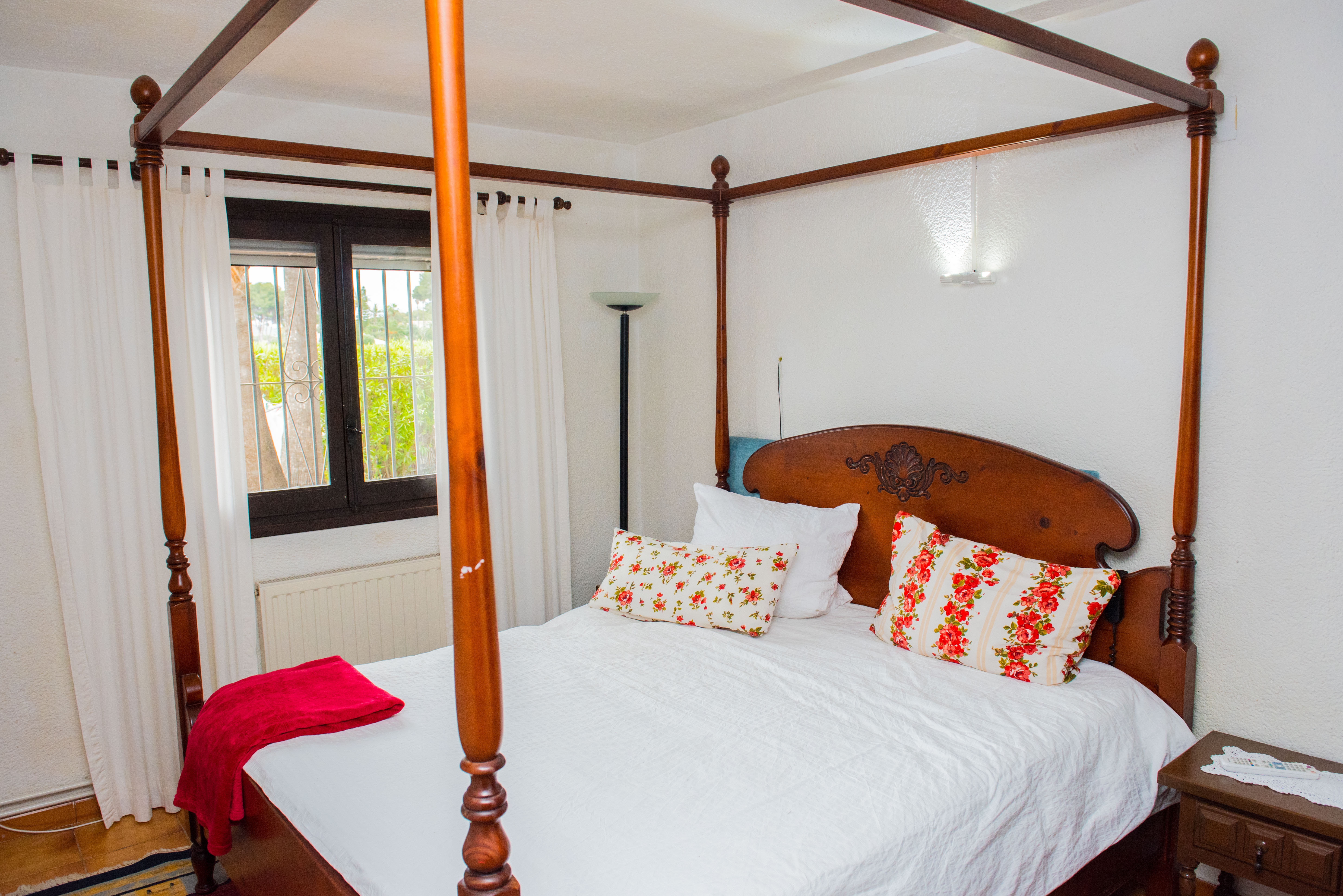 Schlafzimmer mit geschnitztem Himmelbett aus Holz, floralen Kissen und Fenster zum Garten mit darunterliegendem Heizkörper.