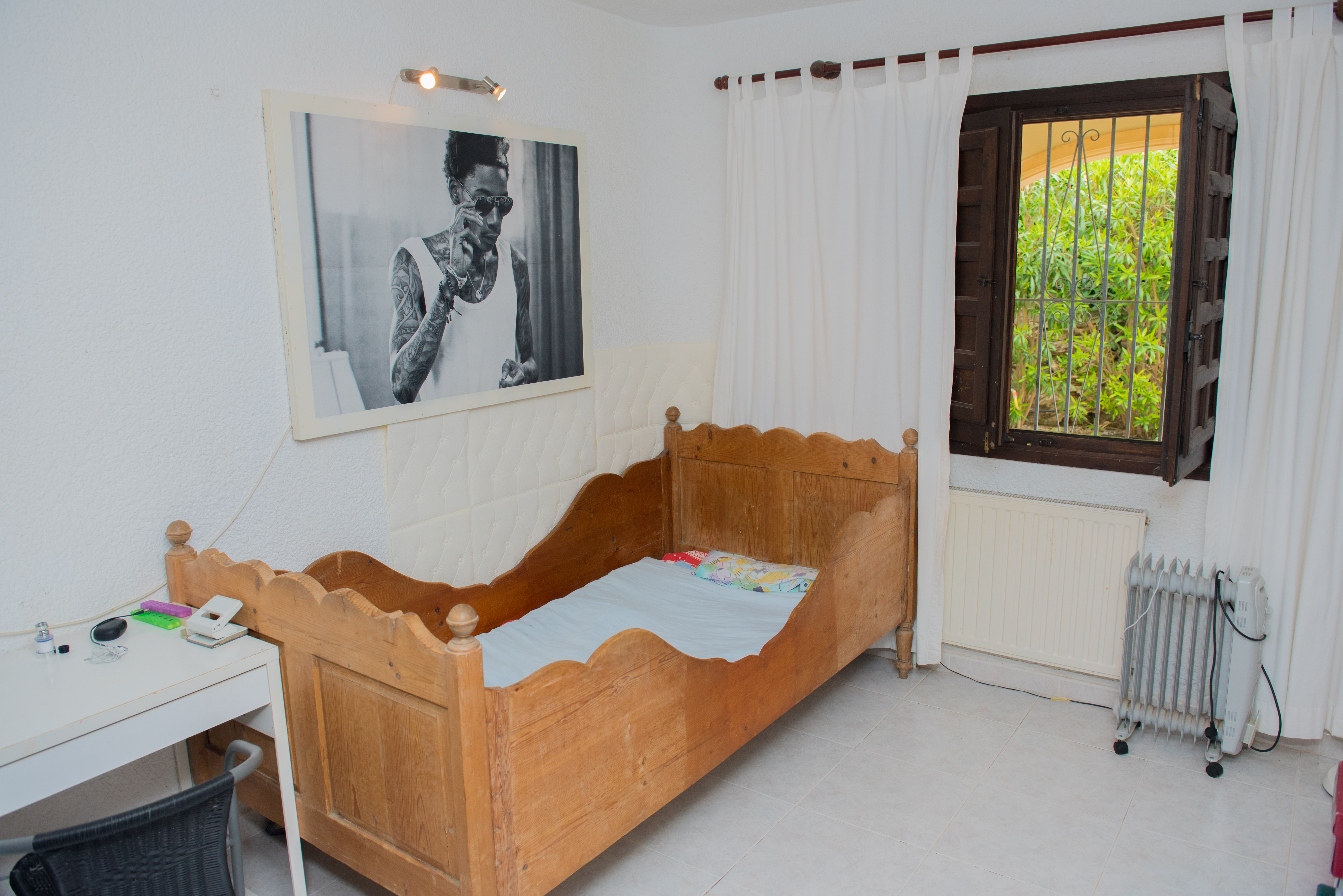 Schlafzimmer mit rustikalem Holzbettgestell, weißem Schreibtisch und dunklem Holzfenster mit Gittern. Heller Fliesenboden und Blick in den Garten.