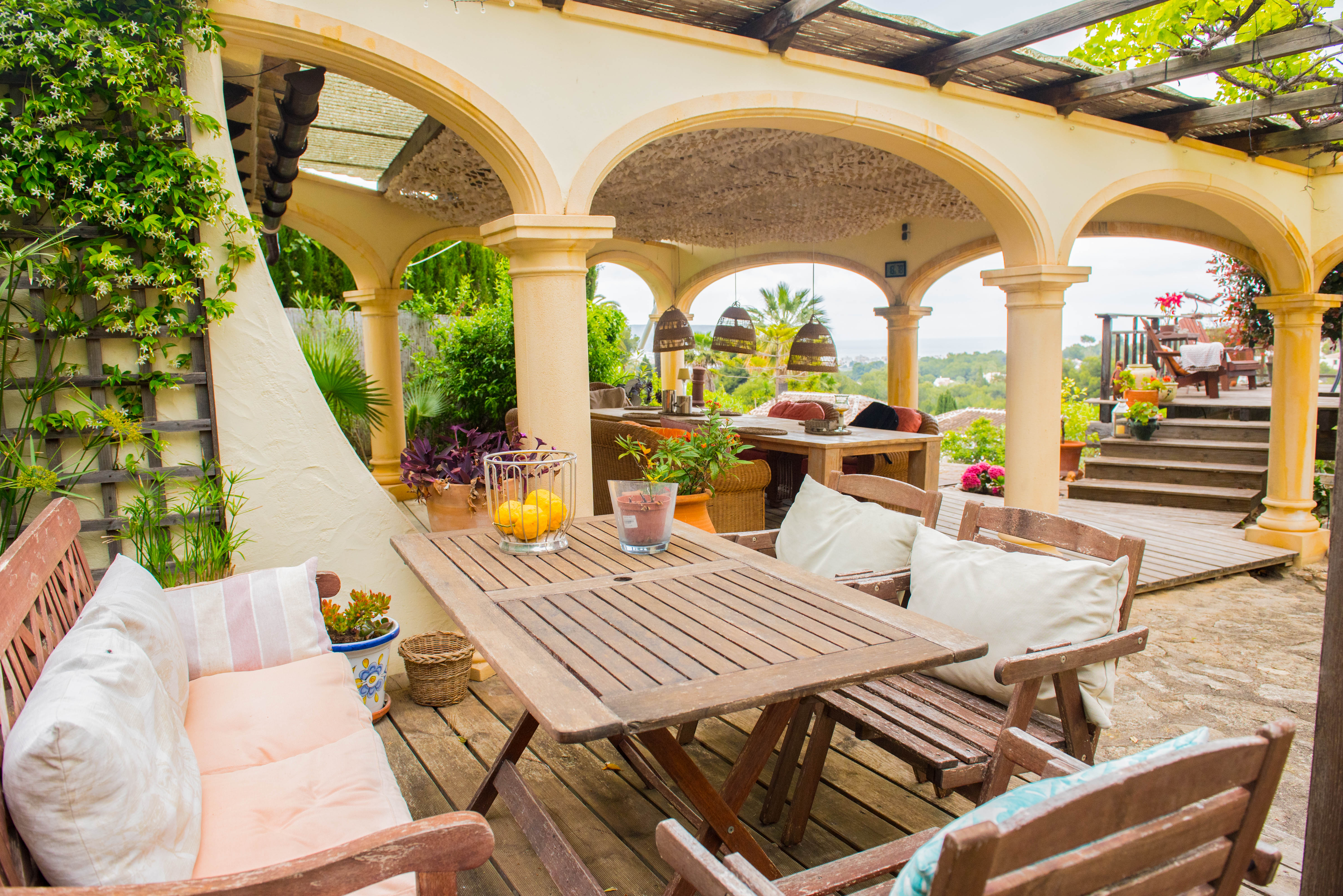 Überdachte Terrasse mit mediterranen Rundbögen, Essbereich aus Holz, Holzdeck und Panoramablick auf das Meer und die grüne Umgebung.