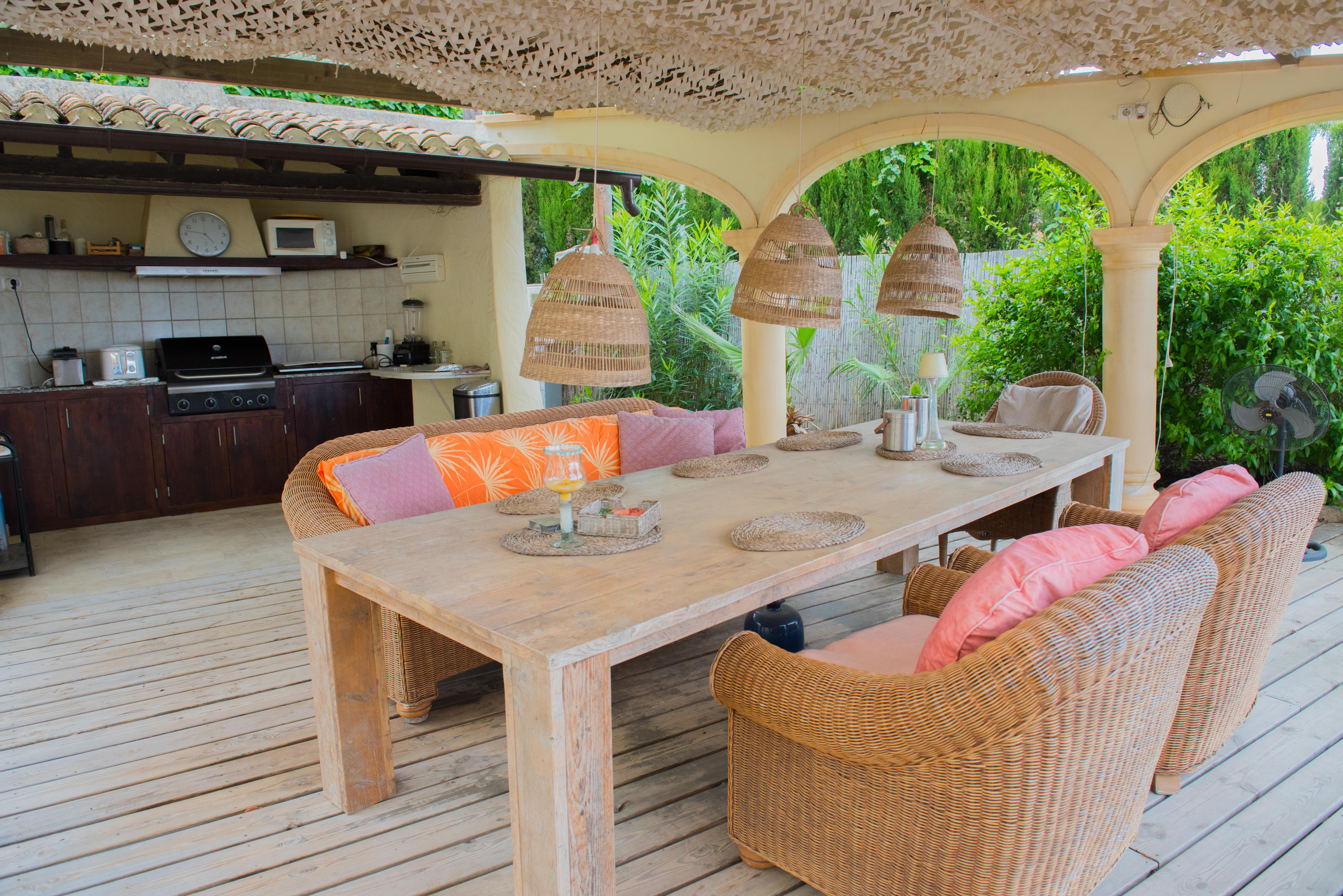 Überdachte Terrasse mit voll ausgestatteter Sommerküche, massivem Holztisch, Korbsesseln und dekorativer Beschattung auf einem Holzdeck.