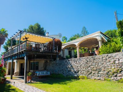 Außenansicht der Villa mit überdachter Holzterrasse und Markise, Garten mit Steinmauer und Ruhebereich.