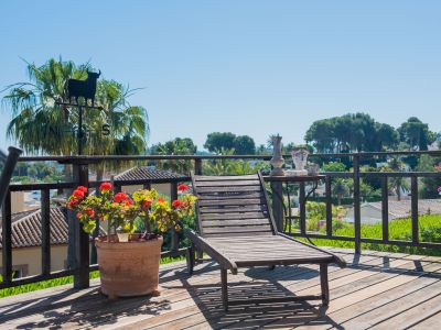 Holzterrasse mit Liegestuhl, Gartenblick und dekorativen Details wie einer Stier-Wetterfahne und Geranientöpfen.