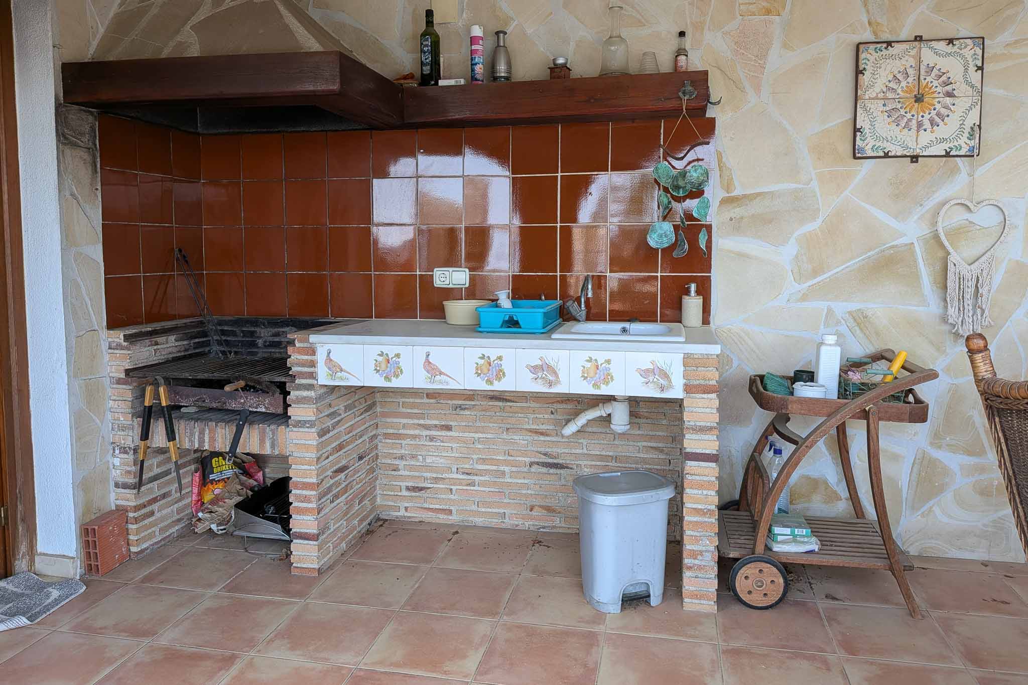 Außen-Sommerküche mit gemauertem Grill, integrierter Spüle und dekorativer Fliesentheke vor einer rustikalen Natursteinwand.