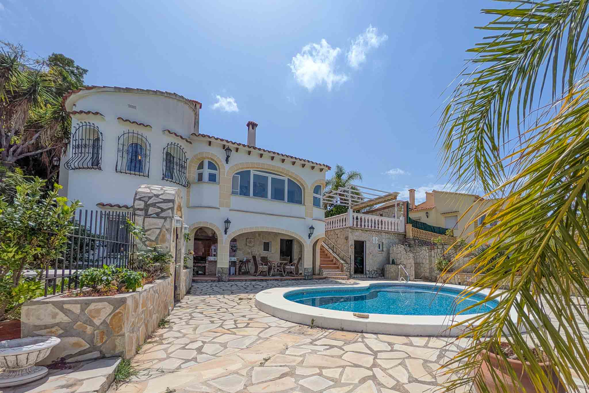 Außenansicht der Villa mit geschwungenem Pool, Natursteinterrasse und überdachter Sommerküche. Die weiße Fassade besticht durch Rundbögen und einen markanten Turmanbau.