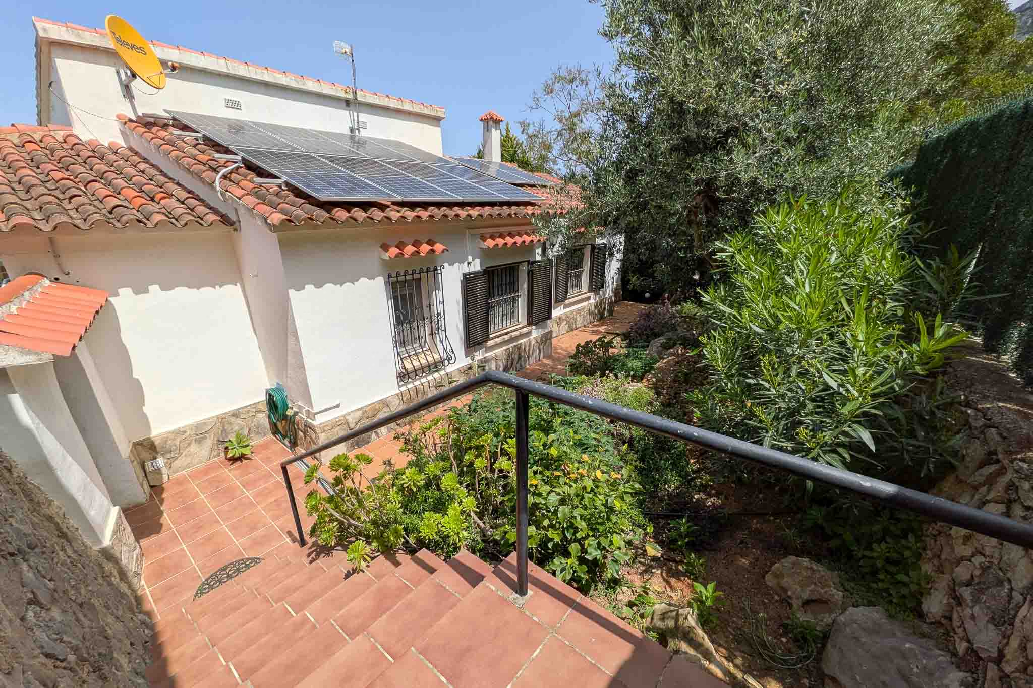 Außenansicht der weißen Fassade mit Photovoltaikanlage auf dem Ziegeldach, Terrakotta-Treppen und gepflegter mediterraner Bepflanzung.
