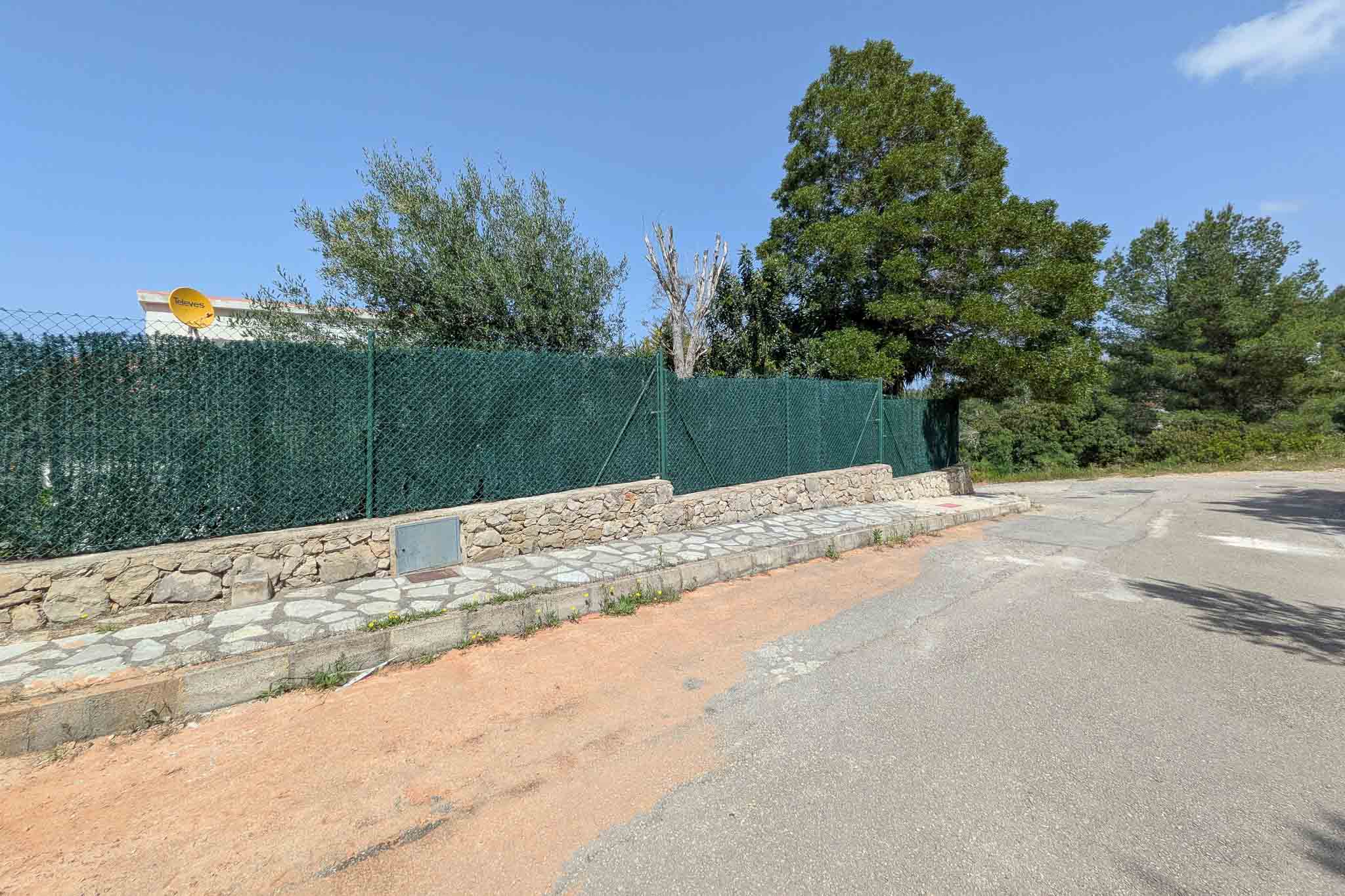Außenansicht mit Natursteinmauer, grünem Sichtschutzzaun und gepflastertem Gehweg in einer ruhigen Wohnstraße.