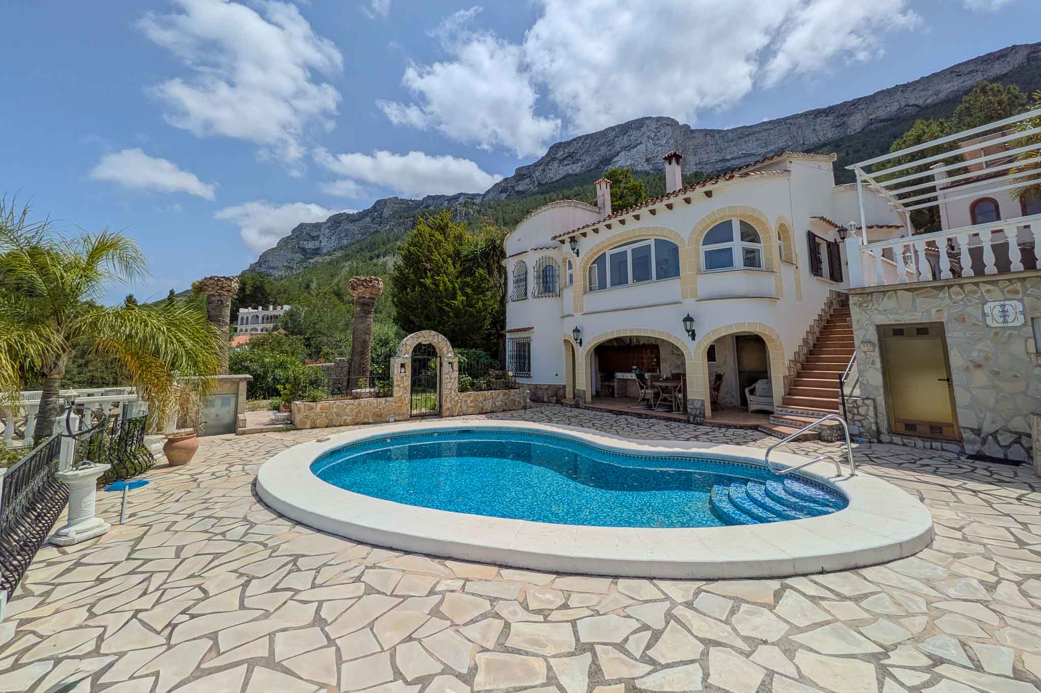 Außenansicht mit nierenförmigem Pool und Natursteinterrasse vor der weißen Fassade mit Rundbögen, im Hintergrund das Montgó-Massiv.
