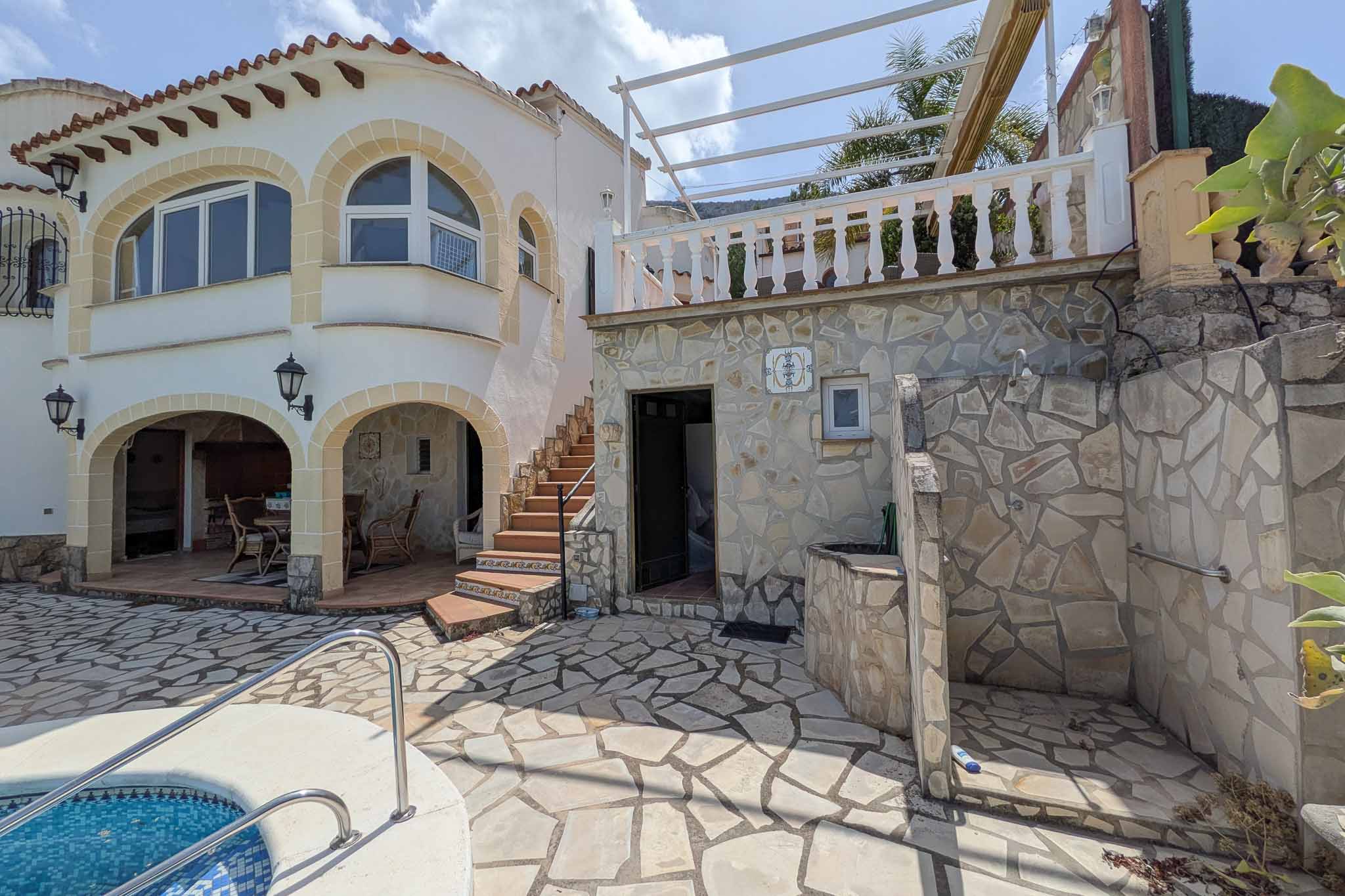 Außenbereich mit Natursteinpflaster, integrierter Außendusche und Treppe zur oberen Terrasse mit weißer Balustrade und Pergola-Struktur.