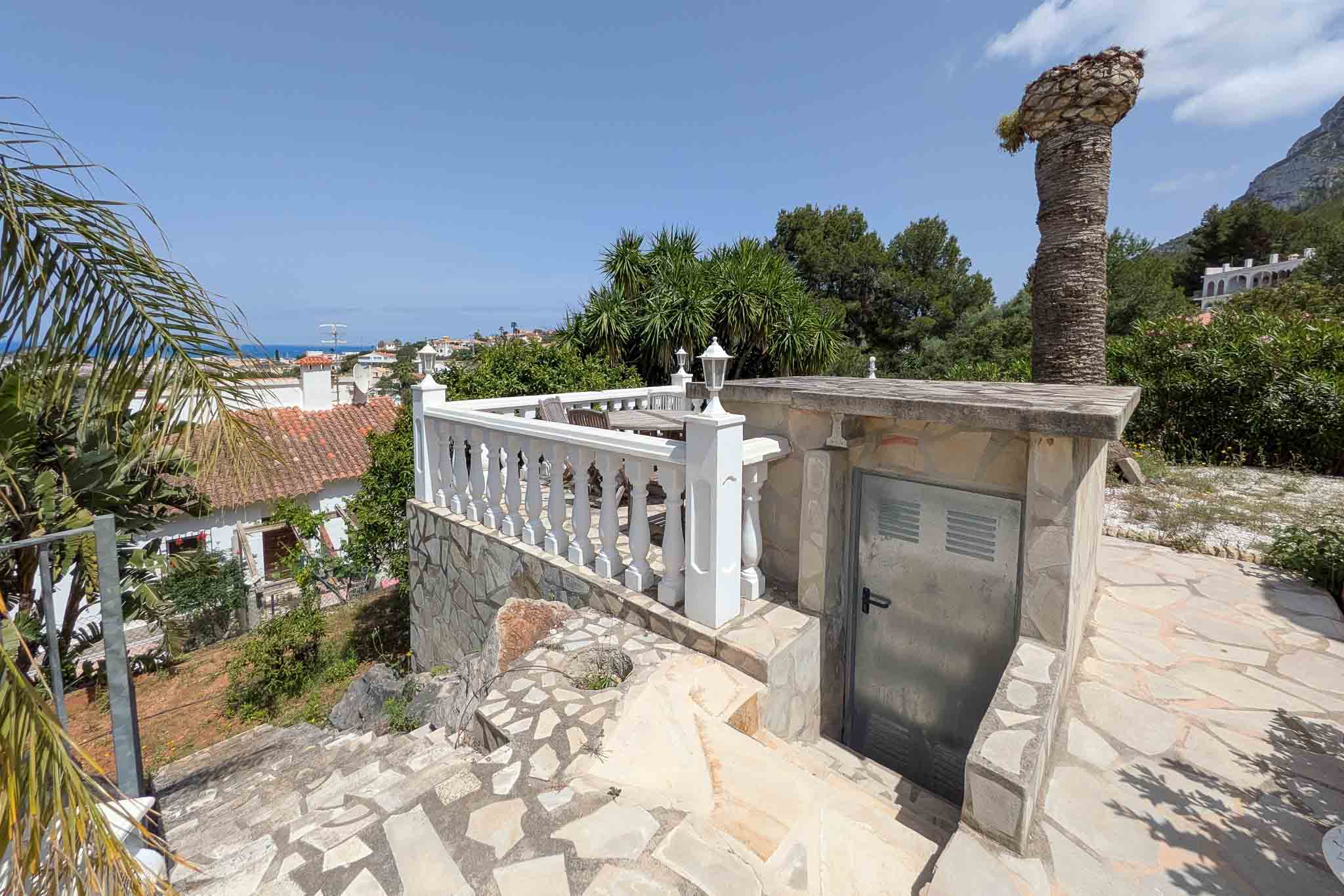Außenterrasse mit weißer Balustrade und Natursteinpflaster, die einen weiten Meerblick und Zugang zu einem technischen Lagerraum bietet.