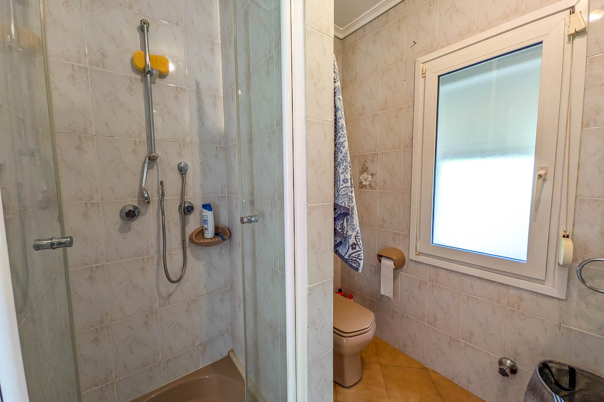Badezimmer mit verglaster Duschkabine, cremefarbenem WC und weißem PVC-Fenster für natürliches Licht. Die Wände sind mit dezent gemusterten Keramikfliesen gestaltet.