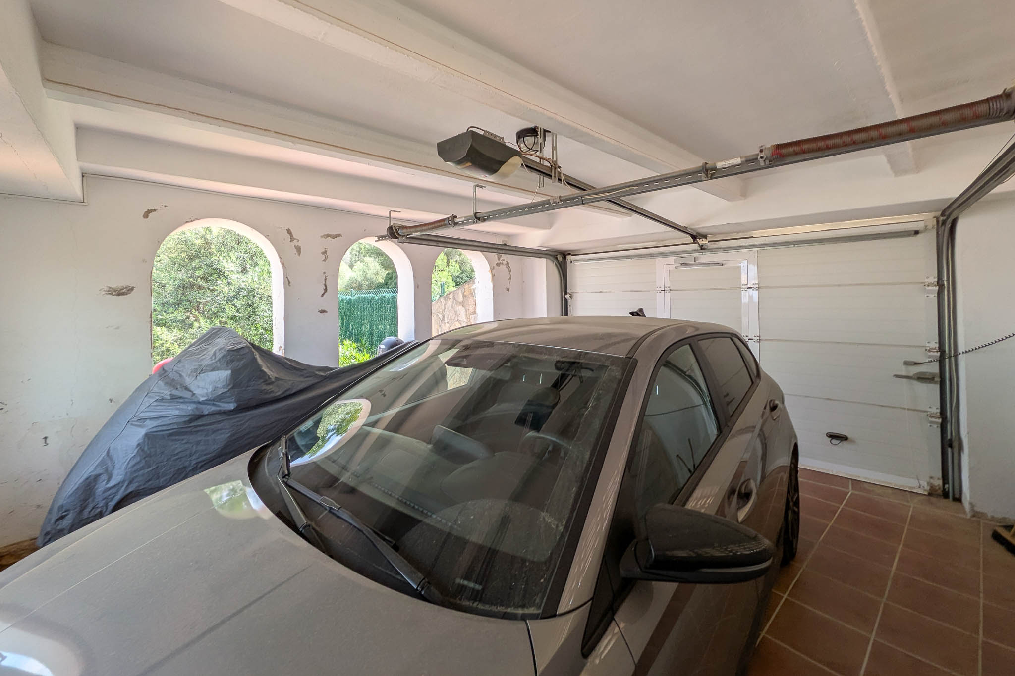 Geschlossene Garage mit automatischem Sektionaltor, Fliesenboden und Deckenbalken. Seitliche Rundbogenöffnungen bieten natürliche Belüftung und Tageslicht.