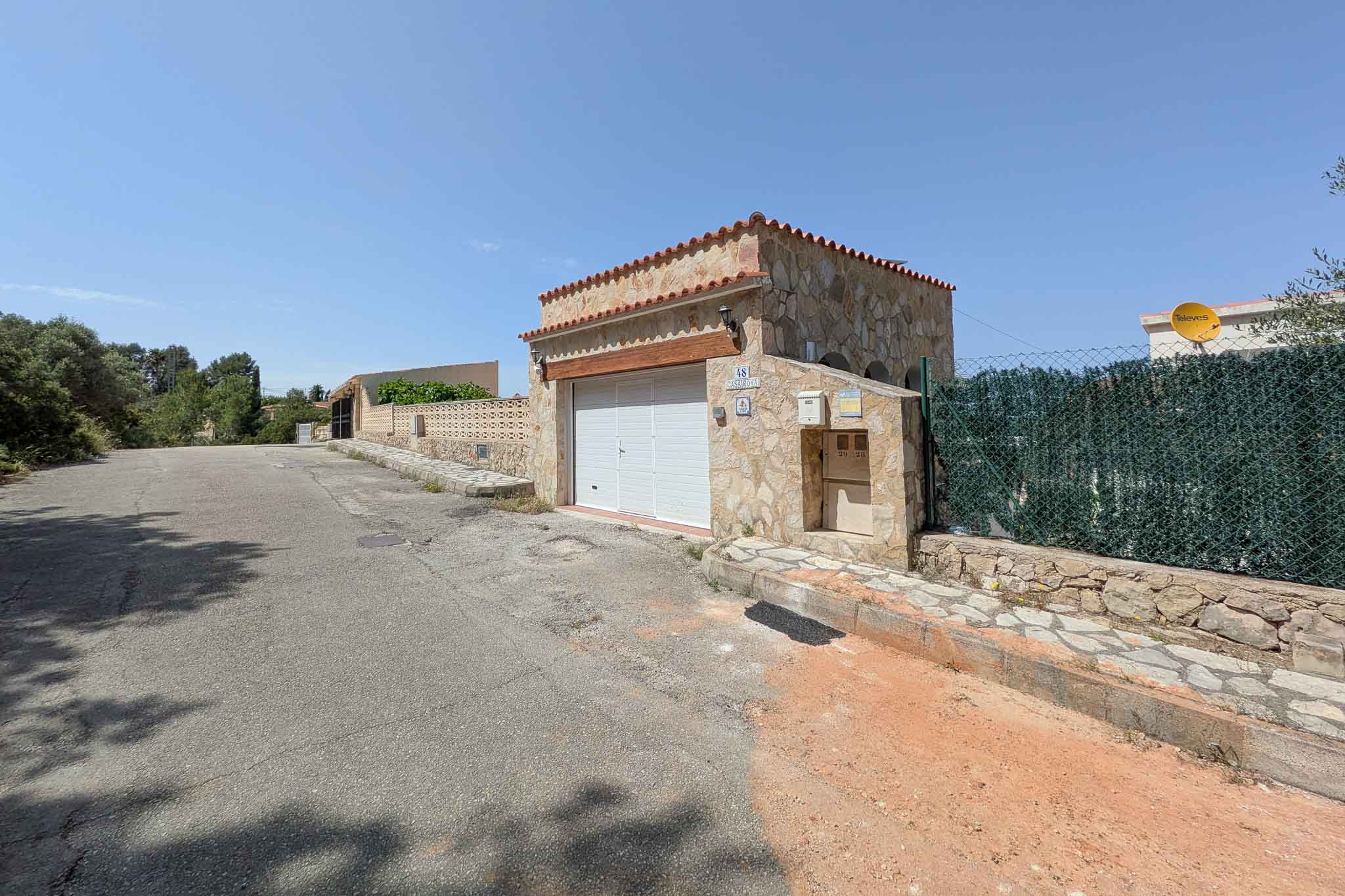 Haupteingang mit einer Garage aus Natursteinmauerwerk, Holzsturz und Ziegeldach an einer ruhigen Zufahrtsstraße in der Zone La Pedrera.