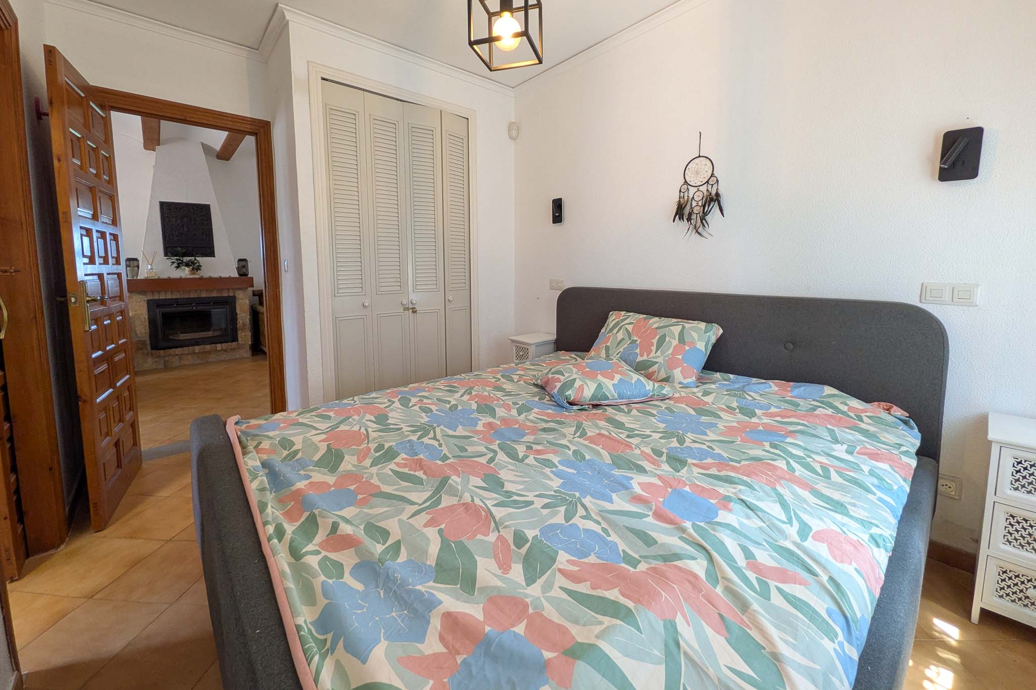 Helles Schlafzimmer mit weißen Einbauschränken im Lamellen-Stil und Blick auf das angrenzende Wohnzimmer mit rustikalem Kamin.