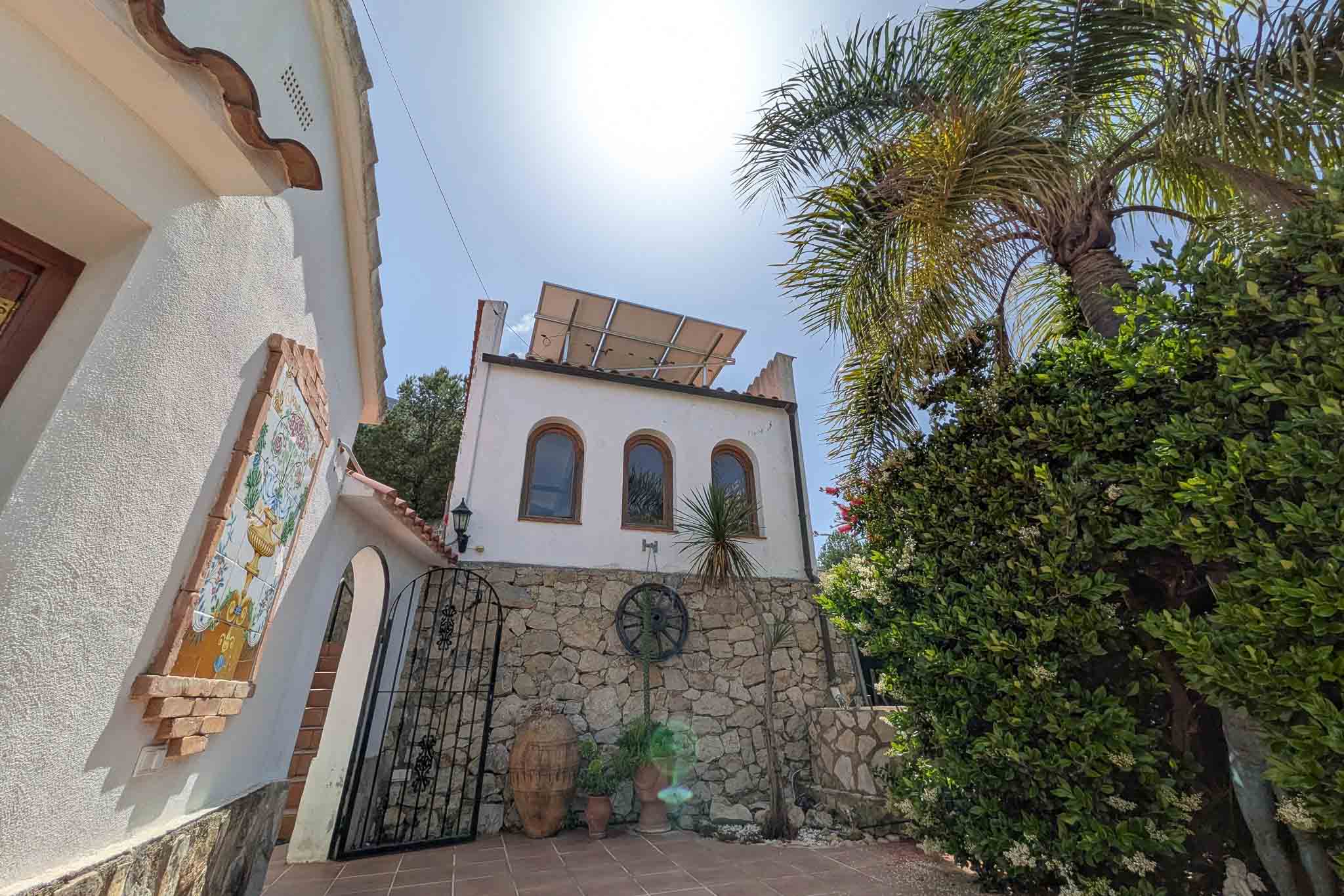 Mediterrane Fassade mit Natursteinmauern, Solaranlage auf dem Dach und traditionellen Details wie einem Keramik-Wandbild und einem schmiedeeisernen Tor.