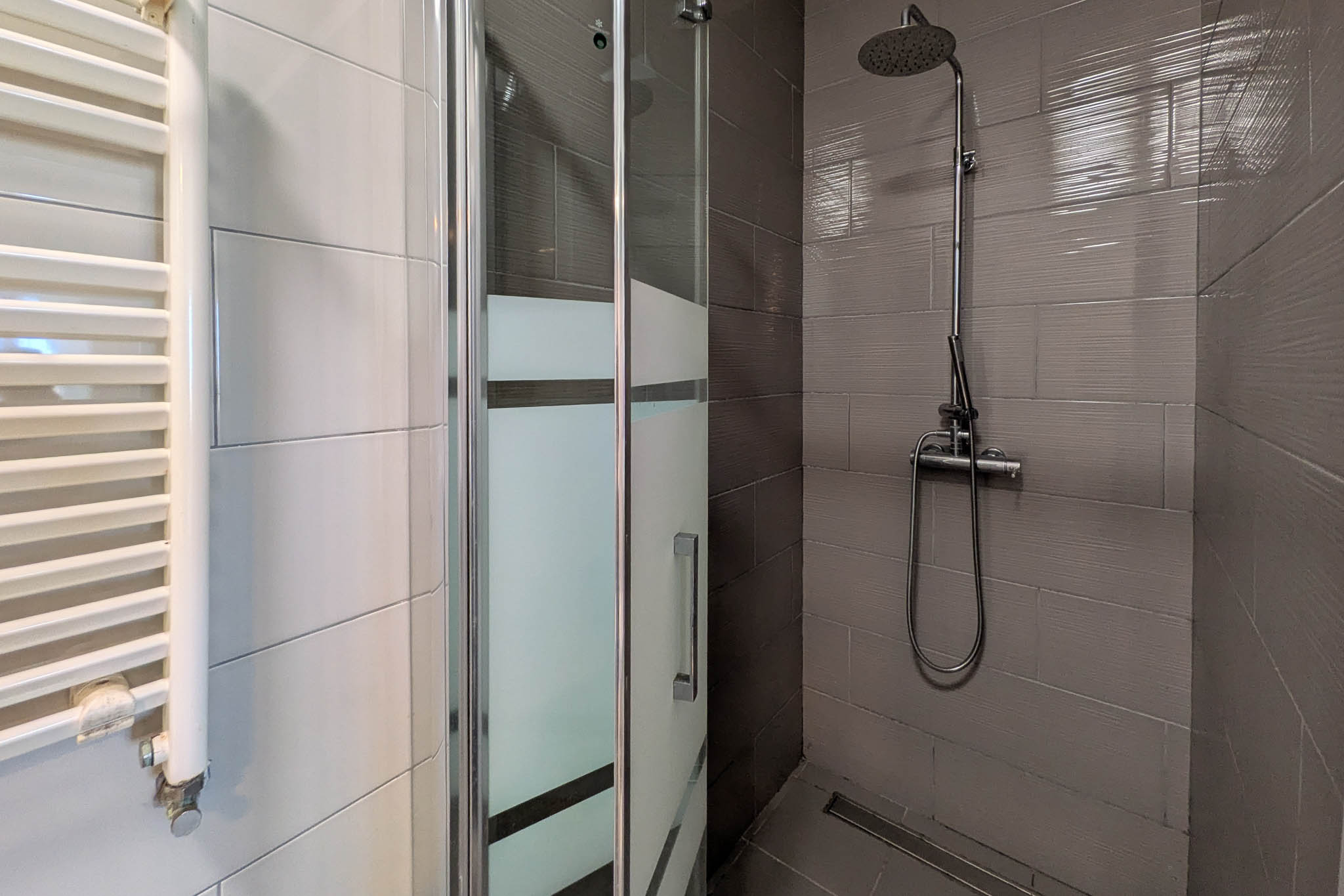 Modernes Badezimmer mit bodengleicher Dusche, Glasabtrennung mit Sichtschutz, verchromten Armaturen und einem weißen Handtuchheizkörper an der Wand.