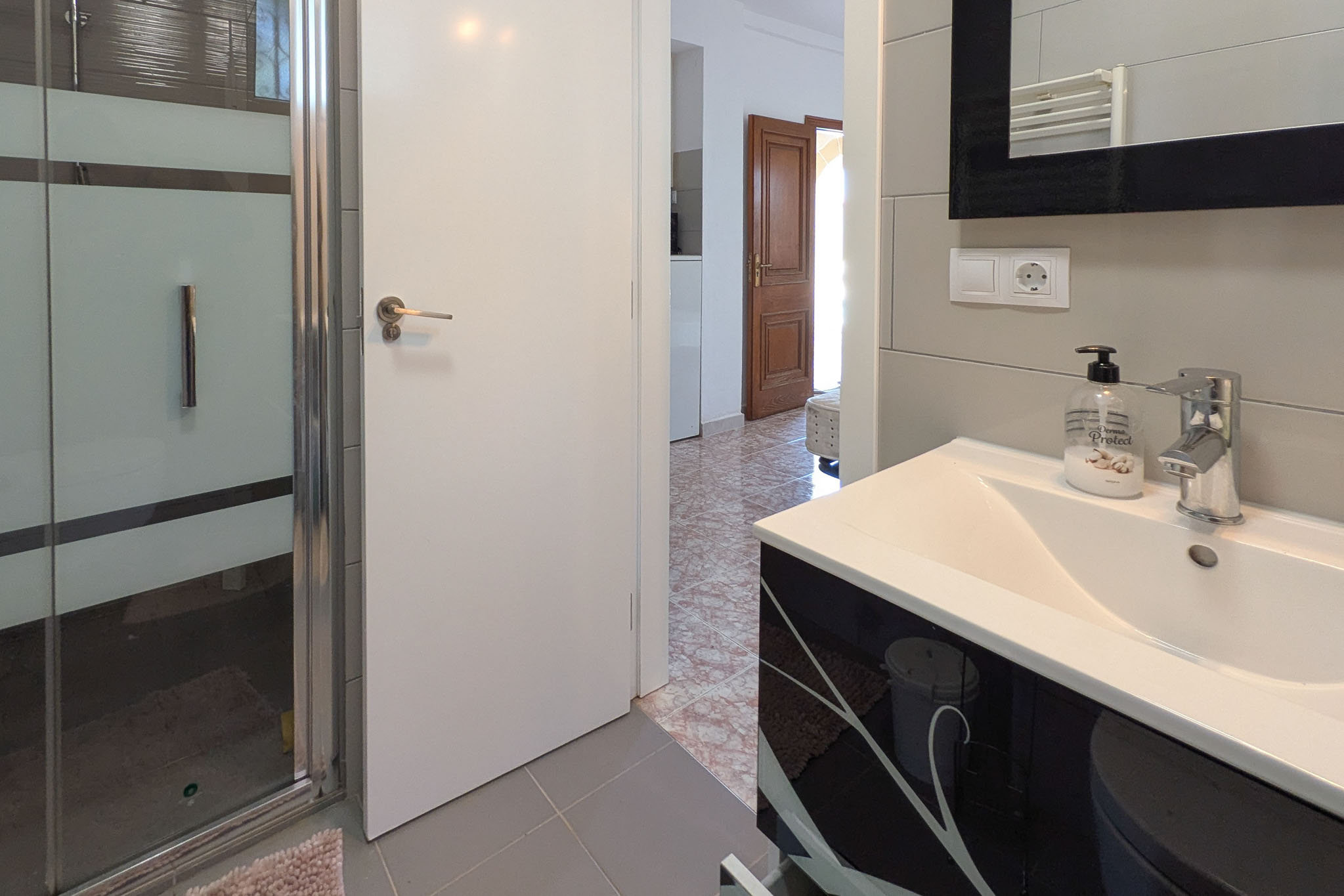Modernes Badezimmer mit gemustertem schwarzem Waschtisch, Glasduschkabine und grauen Fliesen. Der Blick führt durch die weiße Tür in einen Flur mit klassischen Bodenfliesen.
