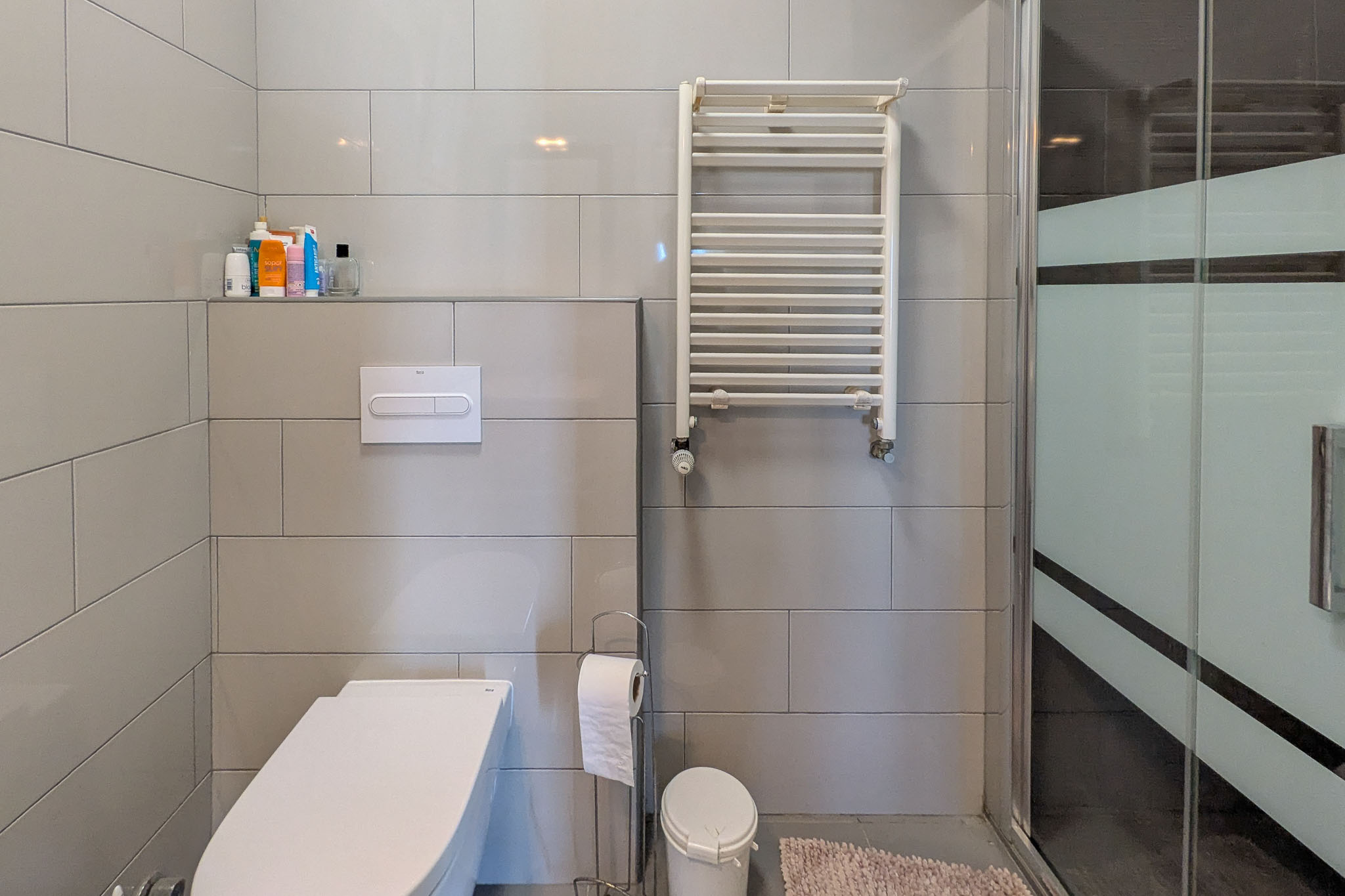 Modernes Badezimmer mit wandhängendem WC, grauen Wandfliesen, einem weißen Handtuchheizkörper und einer Glasduschwand mit Sichtschutzstreifen.