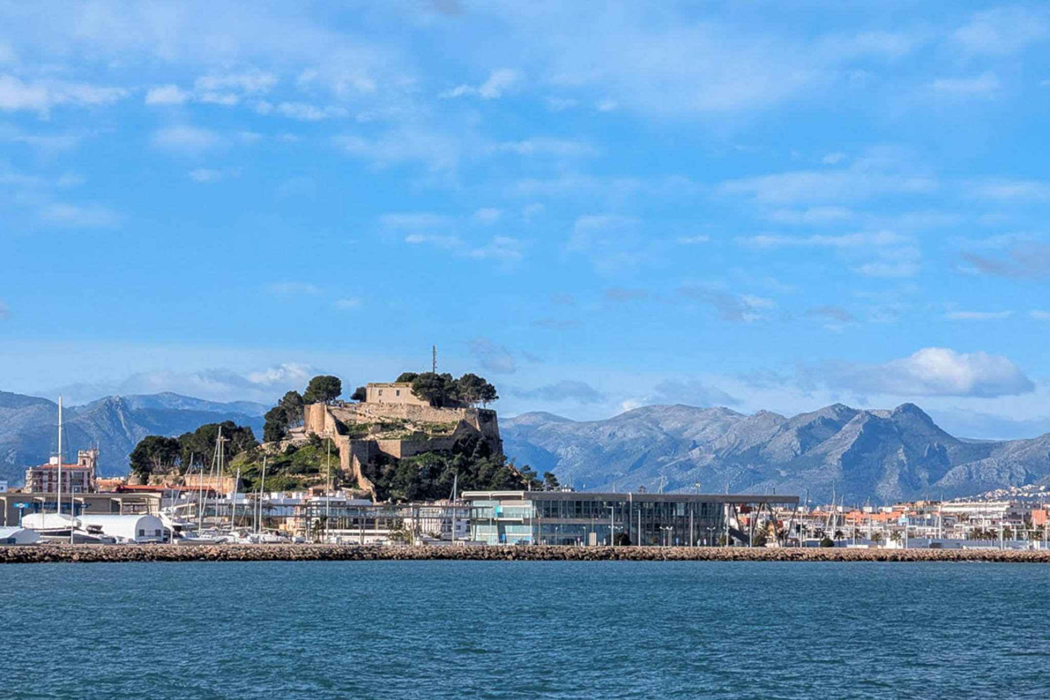 Panoramablick auf die Burg von Dénia und den Hafen, mit der historischen Festung auf dem Hügel und der Bergkette im Hintergrund.