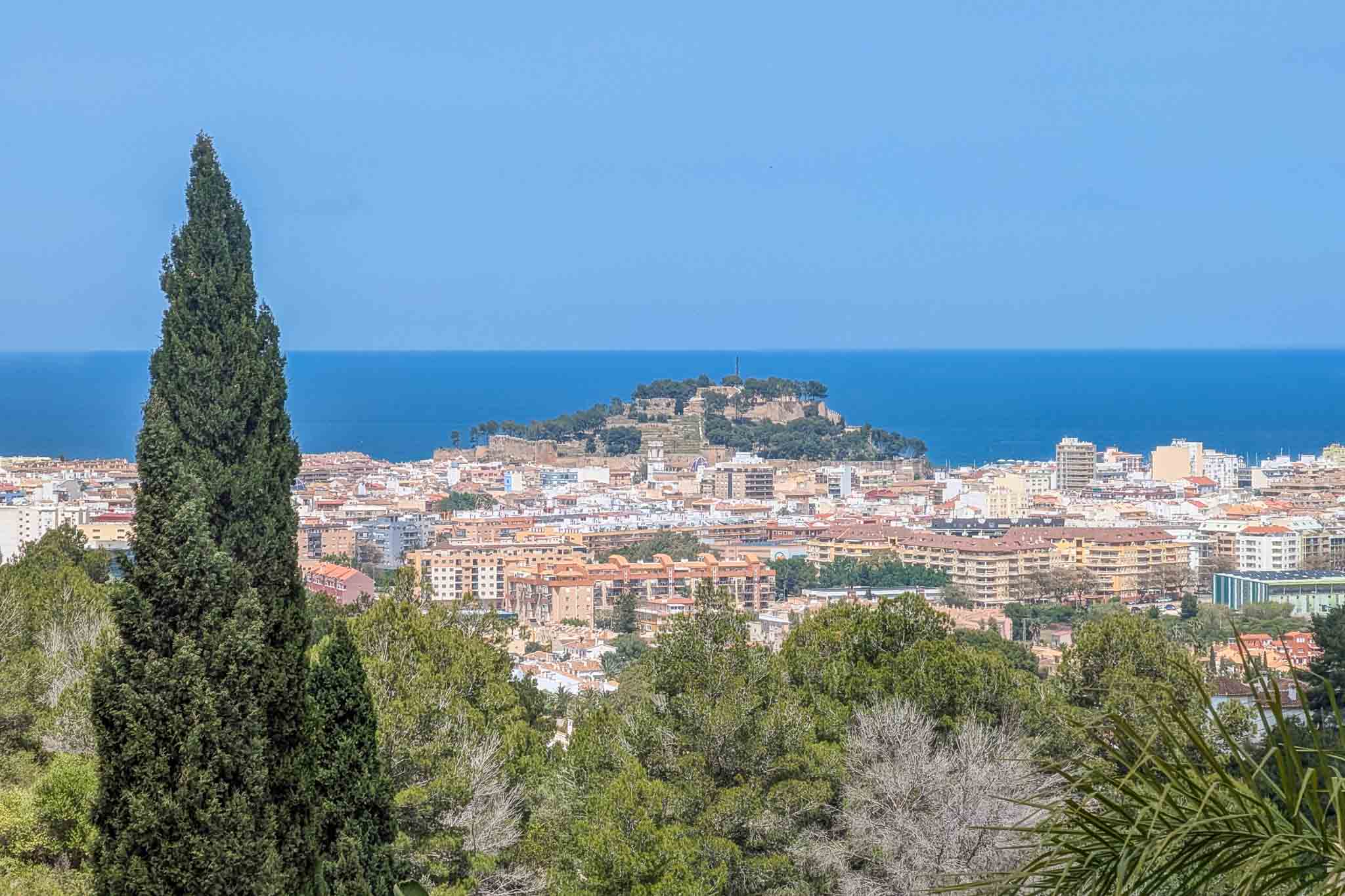 Panoramablick vom Grundstück auf die Burg von Dénia und das Mittelmeer, eingerahmt von mediterraner Vegetation und Zypressen.