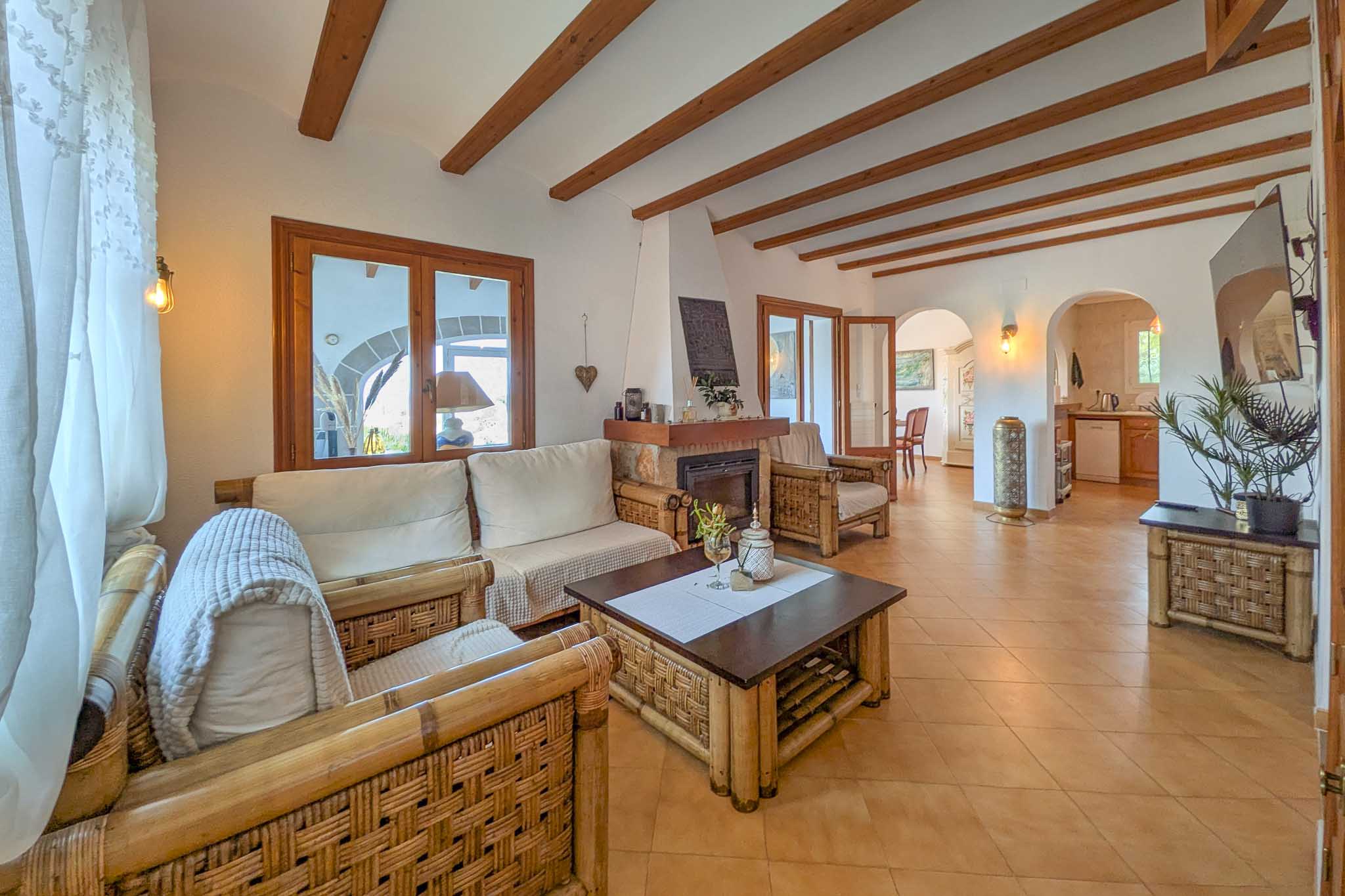 Rustikales Wohnzimmer mit sichtbaren Holzbalken, Steinkamin und Bambusmöbeln. Rundbögen führen zum Essbereich und zur angrenzenden Küche.