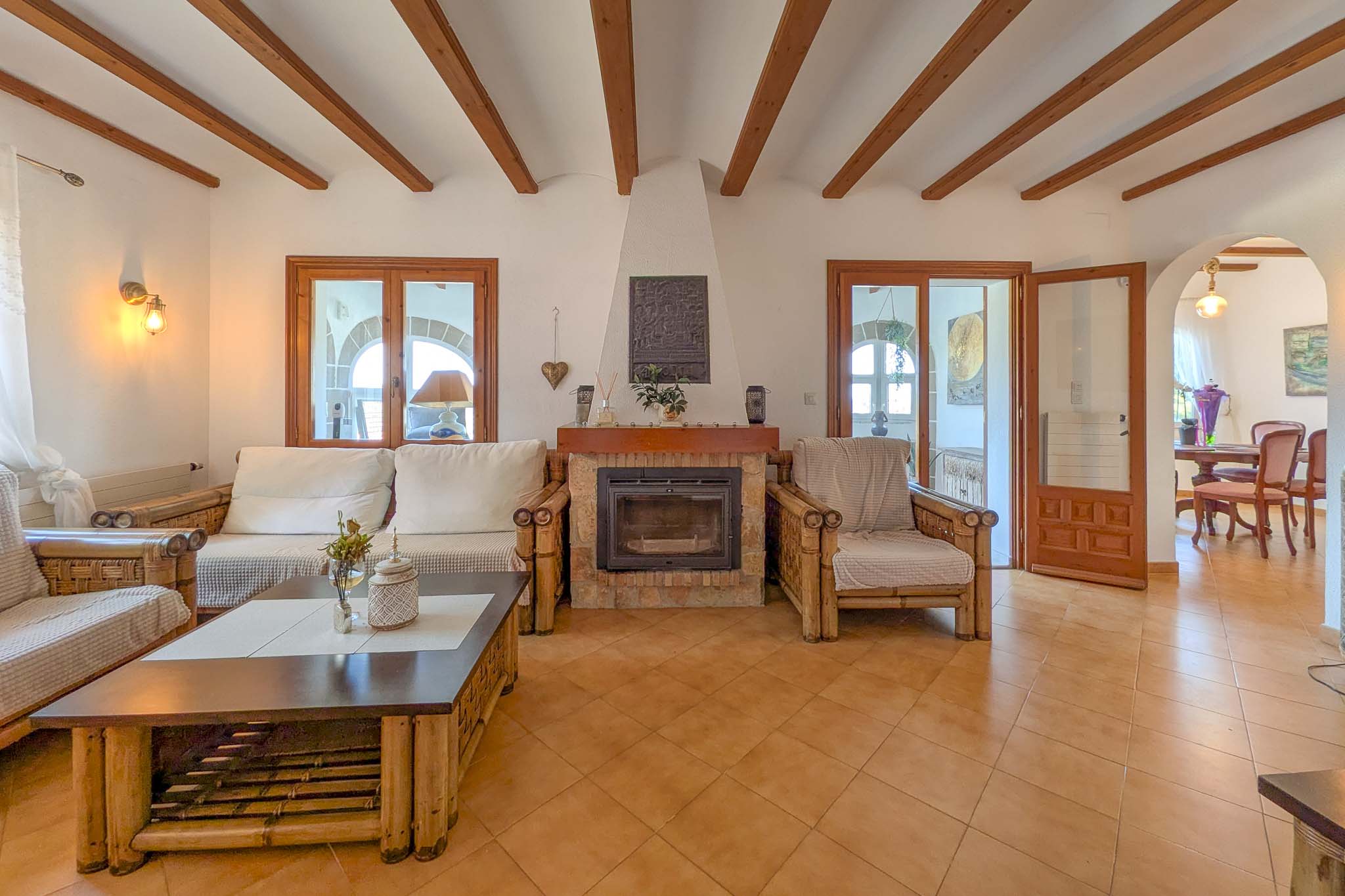 Rustikales Wohnzimmer mit Steinkamin, sichtbaren Holzbalken und Terrakottaboden. Bambusmöbel und Rundbögen zum Essbereich unterstreichen den mediterranen Stil.