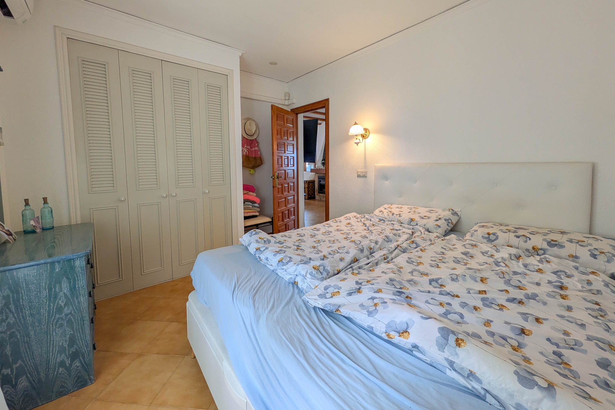 Schlafzimmer mit Einbauschränken im Lamellen-Design, Terrakotta-Fliesen und gepolstertem Kopfteil. Eine blaue Holzkommode setzt Akzente im hellen, neutralen Raum.