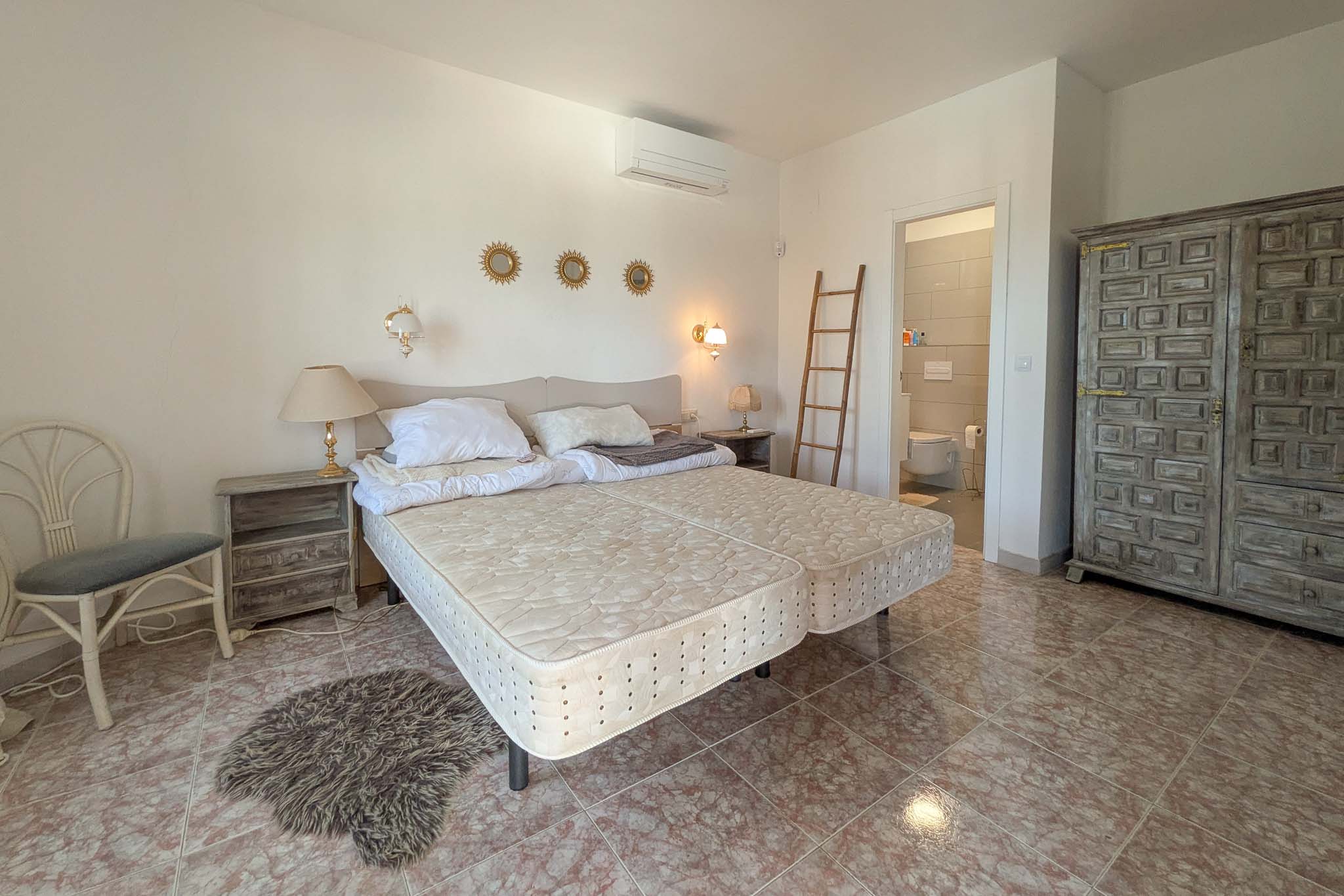Schlafzimmer mit En-Suite-Bad, Fliesenboden in Marmoroptik, rustikalem Schnitzschrank aus Holz und installierter Klimaanlage.