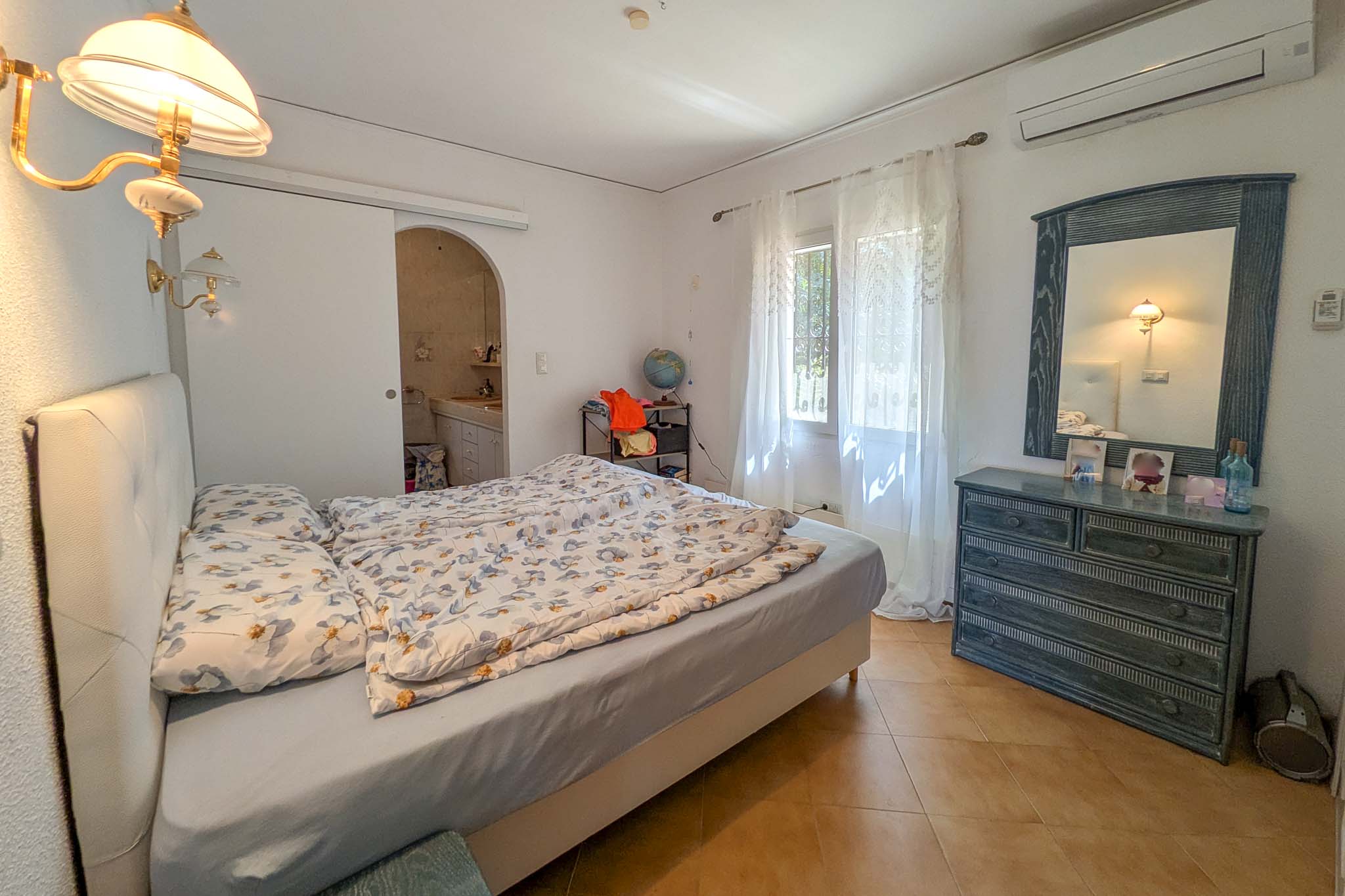 Schlafzimmer mit En-Suite-Badezimmer, Split-Klimaanlage, Terrakotta-Fliesenboden und Holzmöbeln. Der Raum bietet natürliches Licht und klassisches Design.