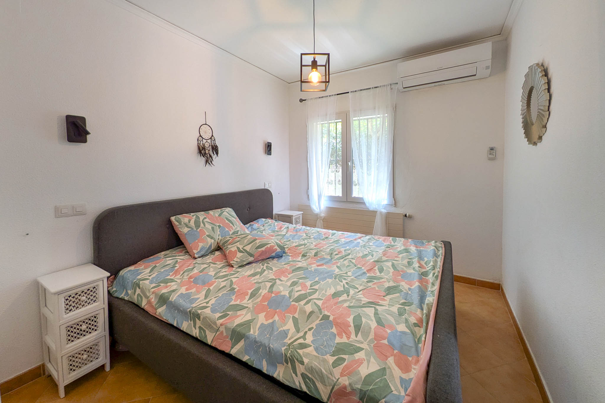 Schlafzimmer mit grau gepolstertem Bettrahmen, Split-Klimaanlage und Terrakotta-Fliesenboden. Der Raum bietet natürliches Licht und eine Hängeleuchte im Industriedesign.