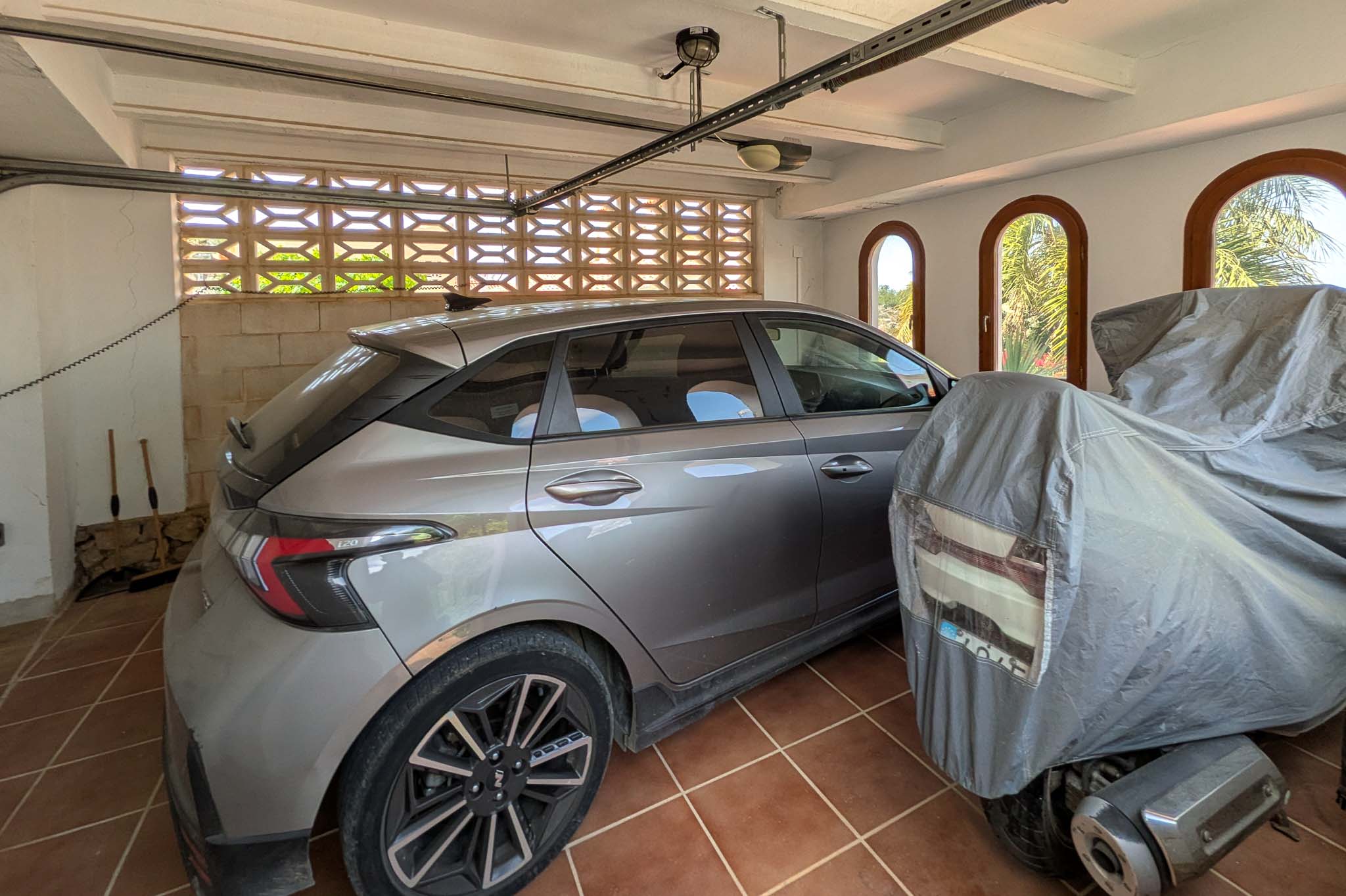 Überdachte Garage mit Terrakottafliesen, weißen Deckenbalken und Rundbogenfenstern. Eine dekorative Gitterwand sorgt für natürliche Belüftung der Stellplätze.