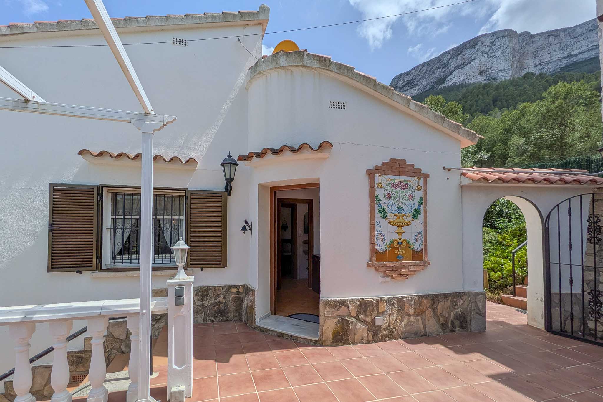 Weiße Fassade mit Natursteinsockel, dekorativem Keramikbild und Terrakottaboden. Im Hintergrund ragt das Montgó-Massiv hinter der bogenförmigen Pforte auf.
