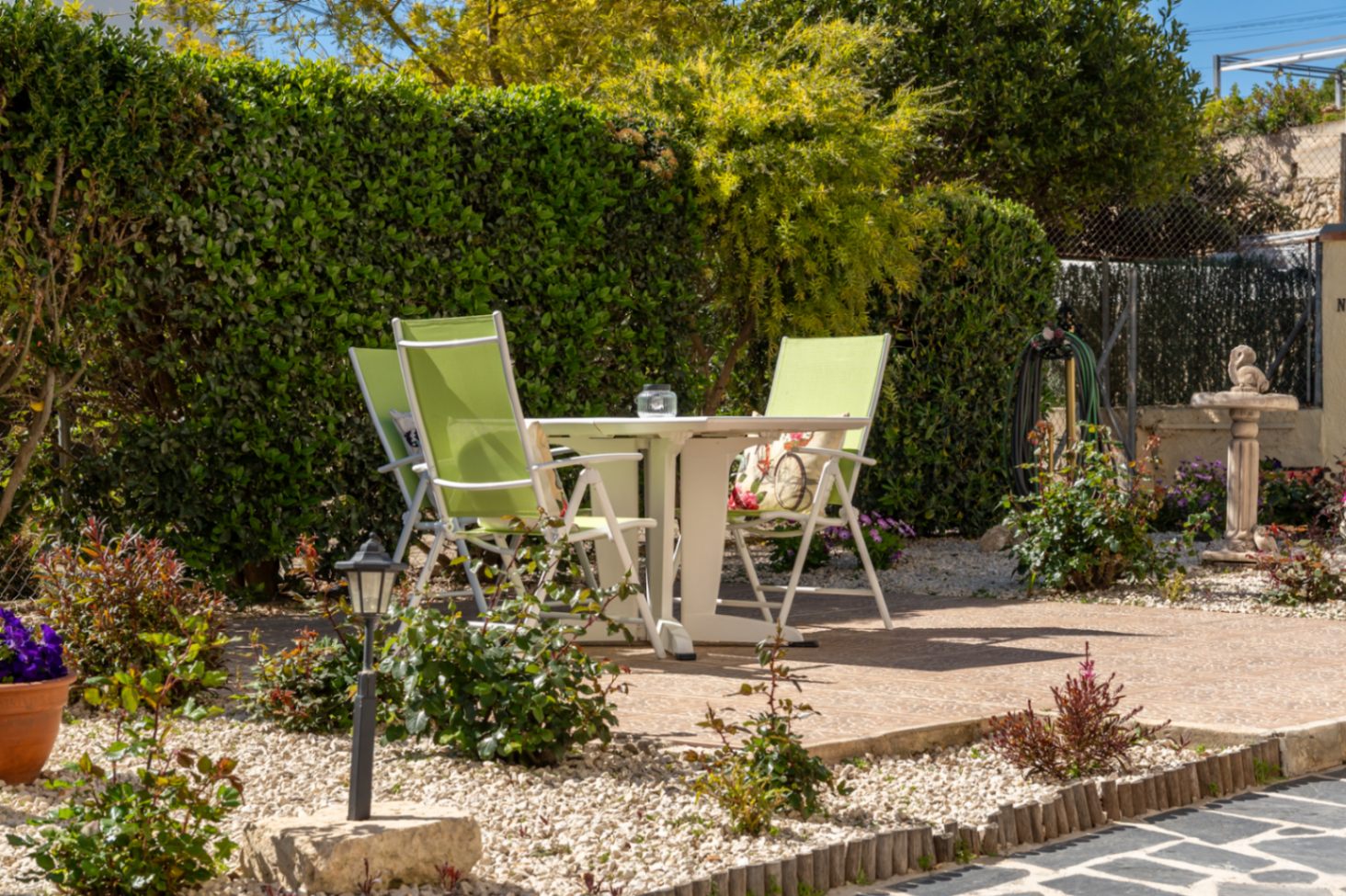 Außenbereich mit weißem Tisch und hellgrünen Stühlen auf einer gepflasterten Terrasse, umgeben von Zierkies und hohen, blickdichten Hecken.
