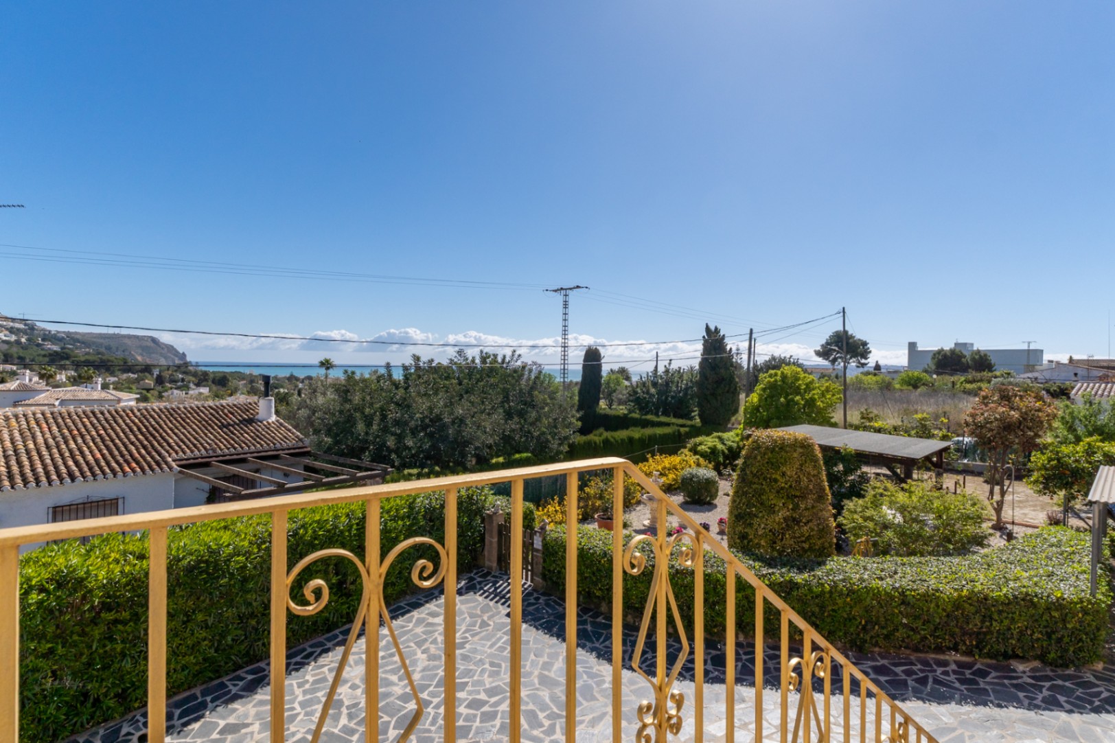 Erhöhter Blick von der Terrasse mit dekorativem Geländer auf den mediterranen Garten und das ferne Meer unter strahlend blauem Himmel.