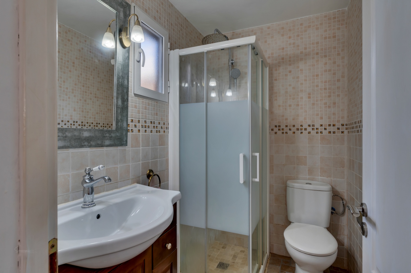 Funktionales Badezimmer mit beige gefliesten Wänden, Duschkabine mit Milchglas, Waschbecken auf Holzunterschrank und Spiegel mit klassischen Wandleuchten.