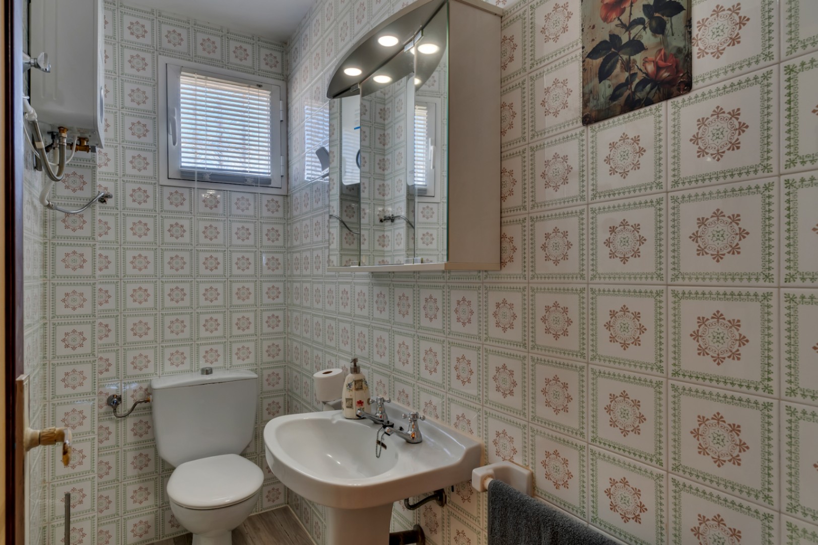 Funktionales Badezimmer mit gemusterten Wandfliesen, Standwaschbecken, WC und Spiegelschrank mit integrierter Beleuchtung.