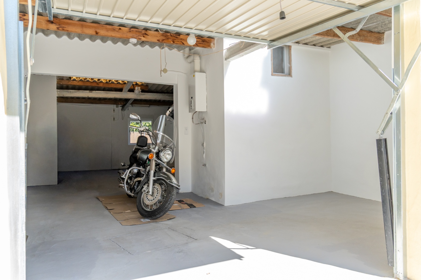 Geschlossene Garage mit Betonboden, weißen Wänden und sichtbaren Holzbalken. Der Raum verfügt über einen wandmontierten Warmwasserbereiter und Platz für Fahrzeuge.