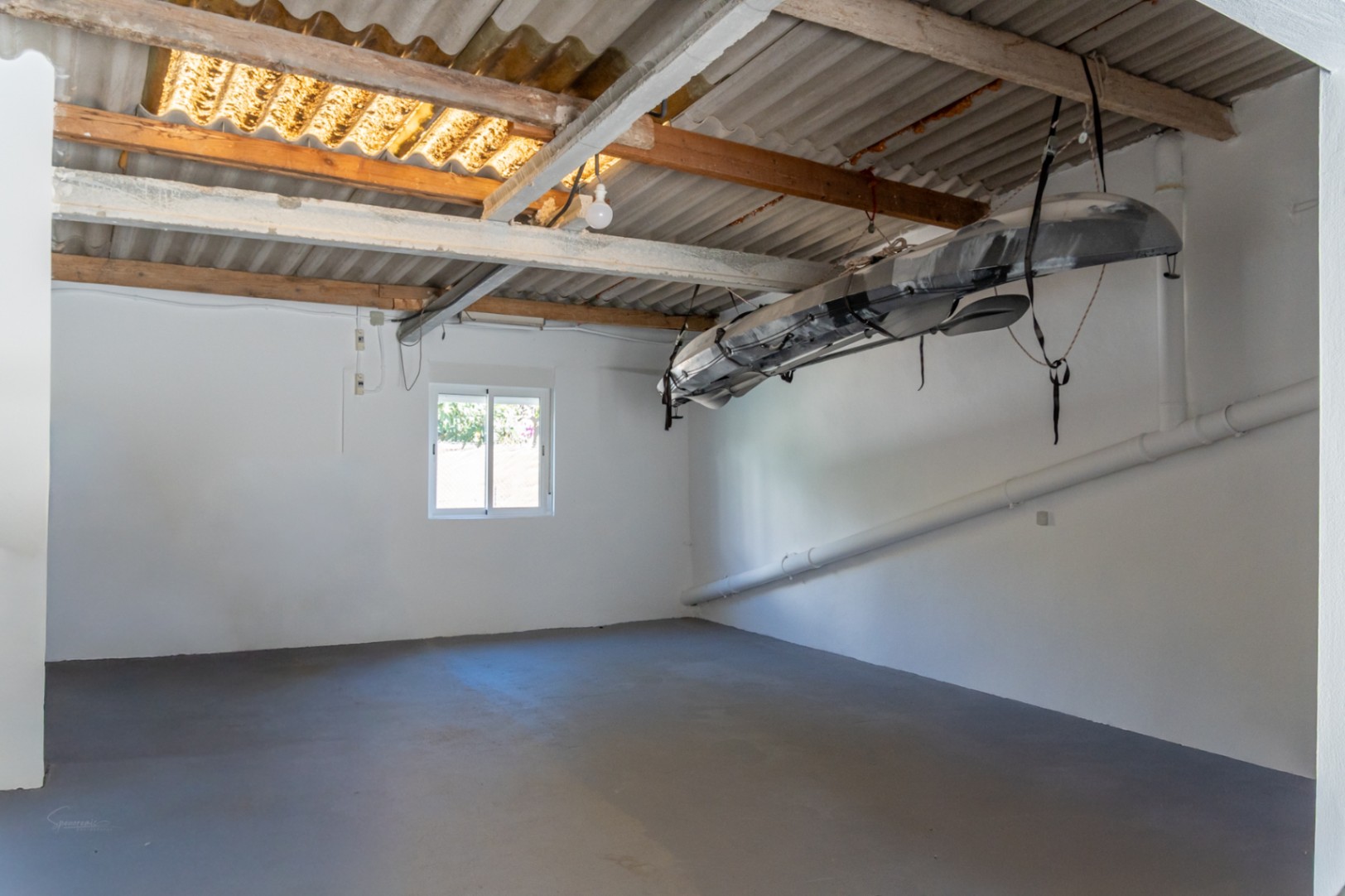 Geschlossene Garage oder Abstellraum mit weißen Wänden, grauem Boden und Deckenhalterung für ein Kajak unter sichtbaren Balken.