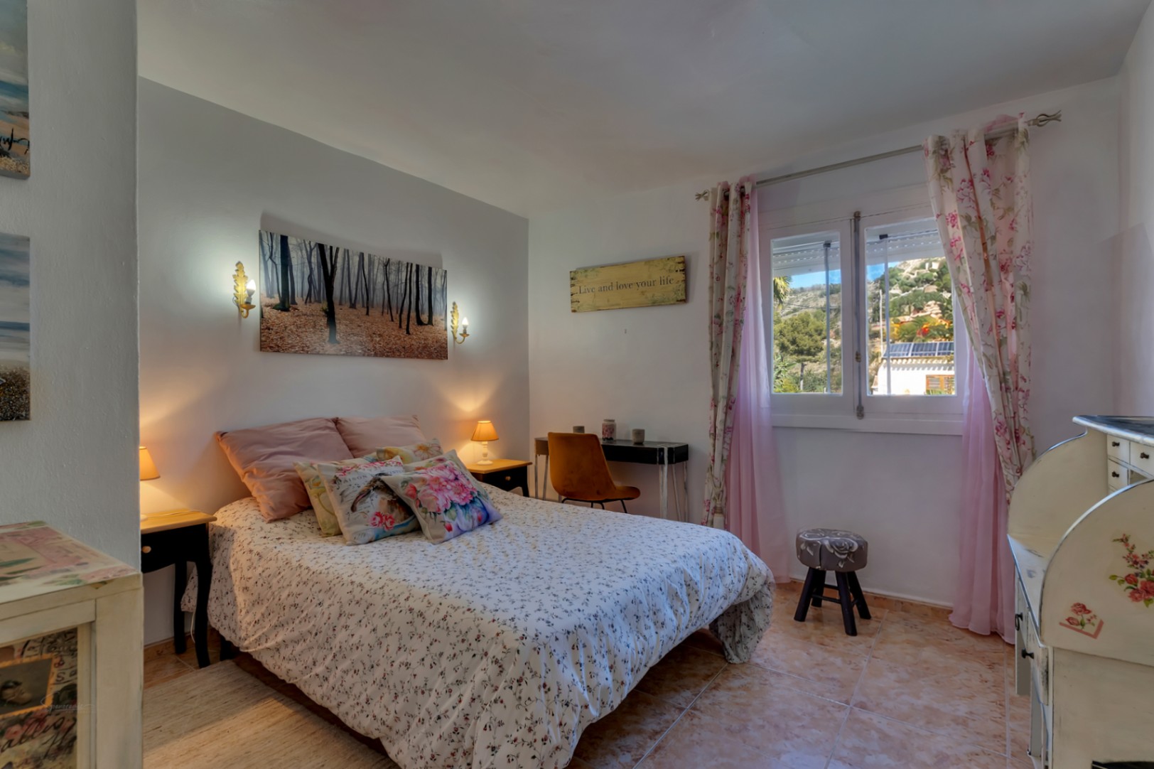 Helles Schlafzimmer mit Fliesenboden, funktionalem Arbeitsplatz und Fenster mit Blick auf die umliegenden Hügel. Klassische Möbel und warme Beleuchtung sorgen für Komfort.