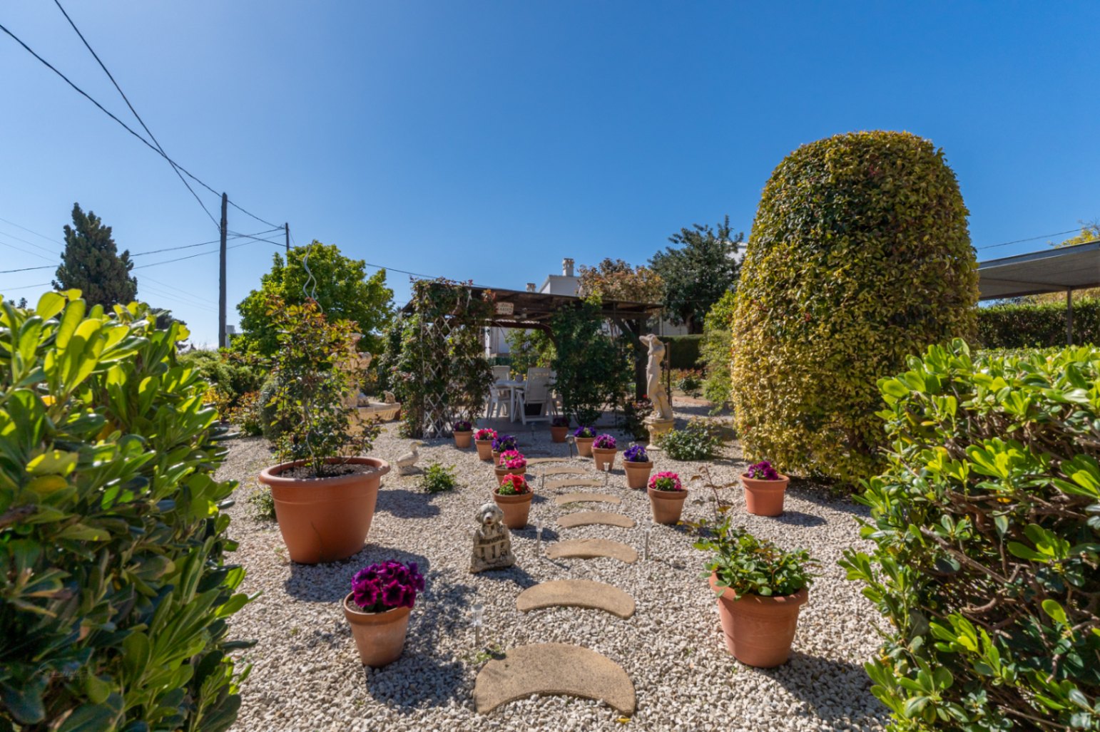 Mediterraner Garten mit weißem Kies, Steinpfad und Terrakotta-Töpfen. Eine Holzpergola bietet einen schattigen Essbereich im Freien inmitten gepflegter Hecken.