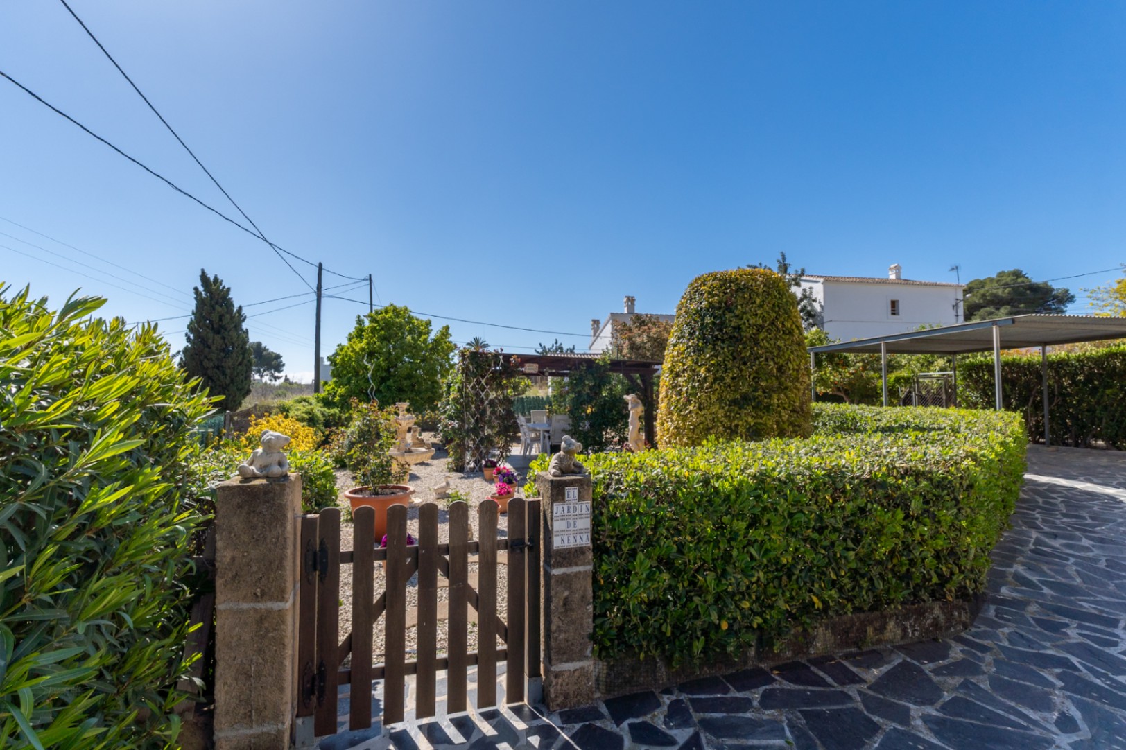 Mediterraner Garteneingang mit Holztor, gepflegten Hecken, Kieswegen und einer schattigen Pergola mit Sitzgelegenheiten im Freien.