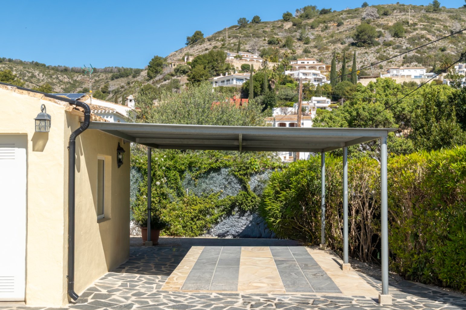 Metall-Carport mit Flachdach und Natursteinpflasterung neben dem Gebäude, mit Blick auf die bewaldeten Hänge von Jávea.