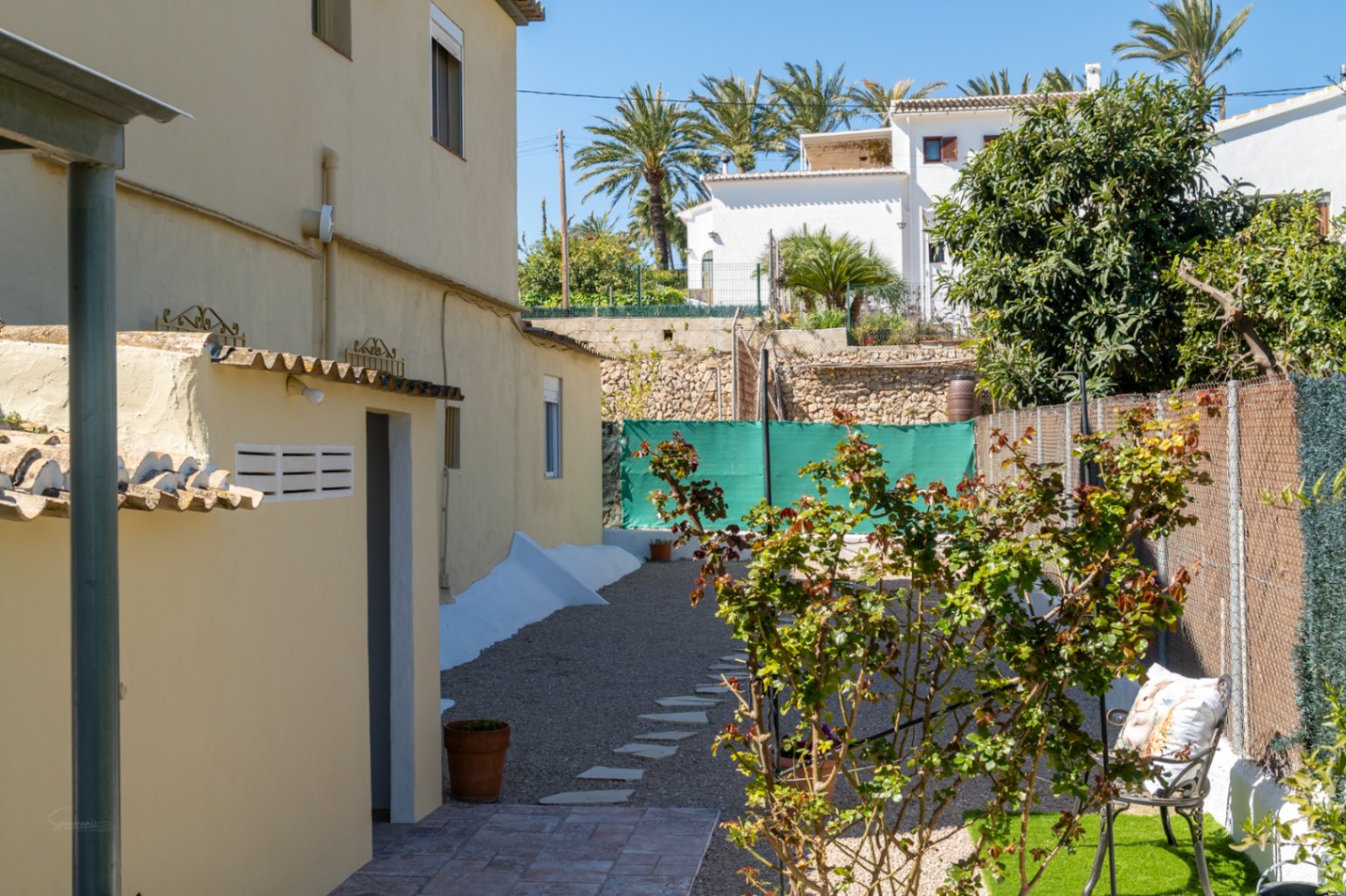 Seitlicher Außenbereich mit Kiesweg, Trittsteinen, mediterraner Bepflanzung und einem funktionalen Nebengebäude auf dem weitläufigen Grundstück.