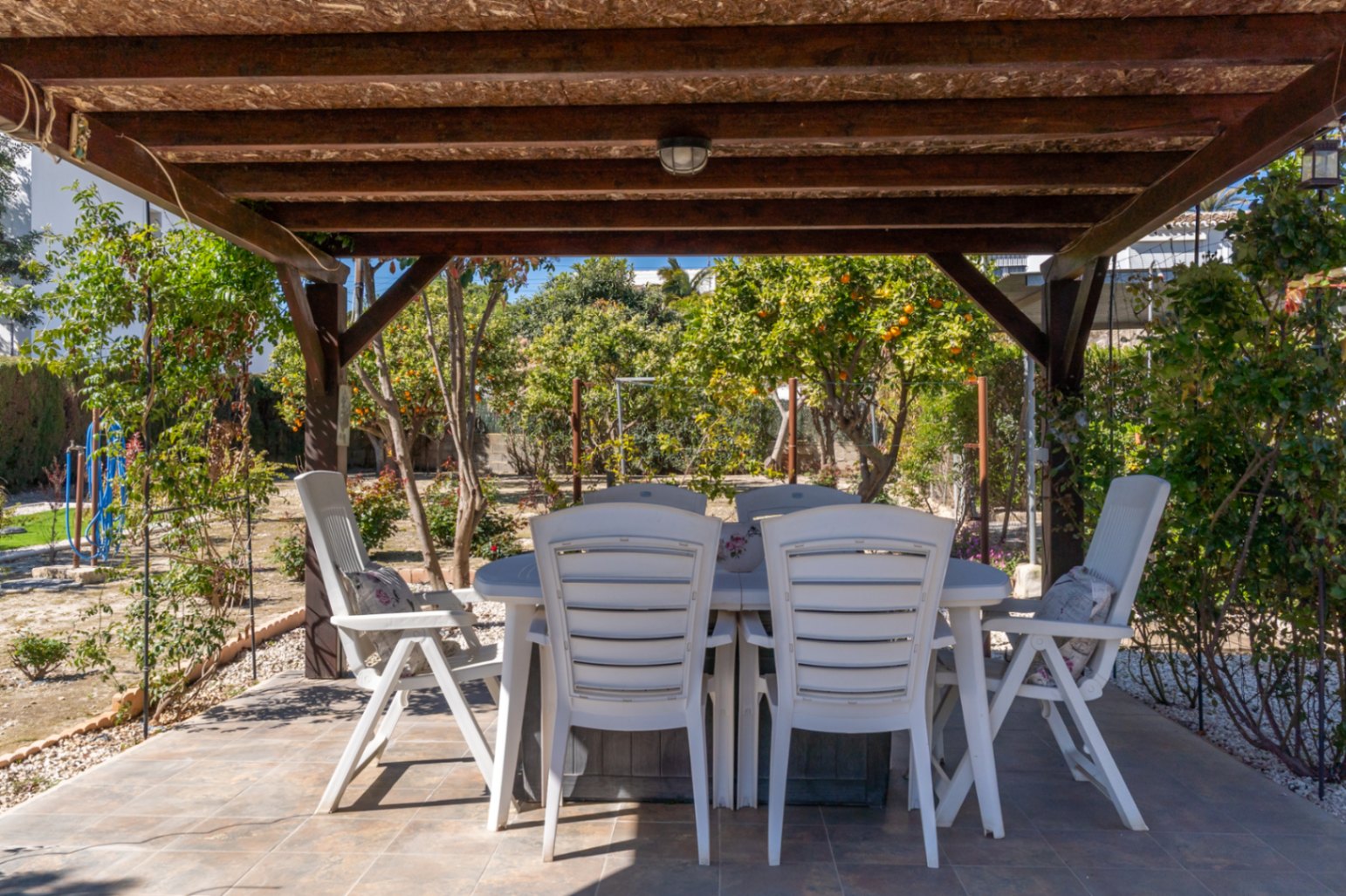 Überdachter Essbereich im Freien mit Holzpergola und weißen Gartenmöbeln, umgeben von einem Garten mit Zitrusbäumen.