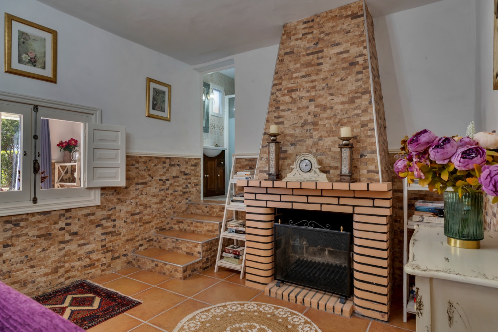 Wohnzimmer mit gemauertem Kamin, Steinverkleidung und Terrakottaboden. Eine kurze Treppe führt zum Badezimmer, ergänzt durch ein Innenfenster mit weißen Fensterläden.