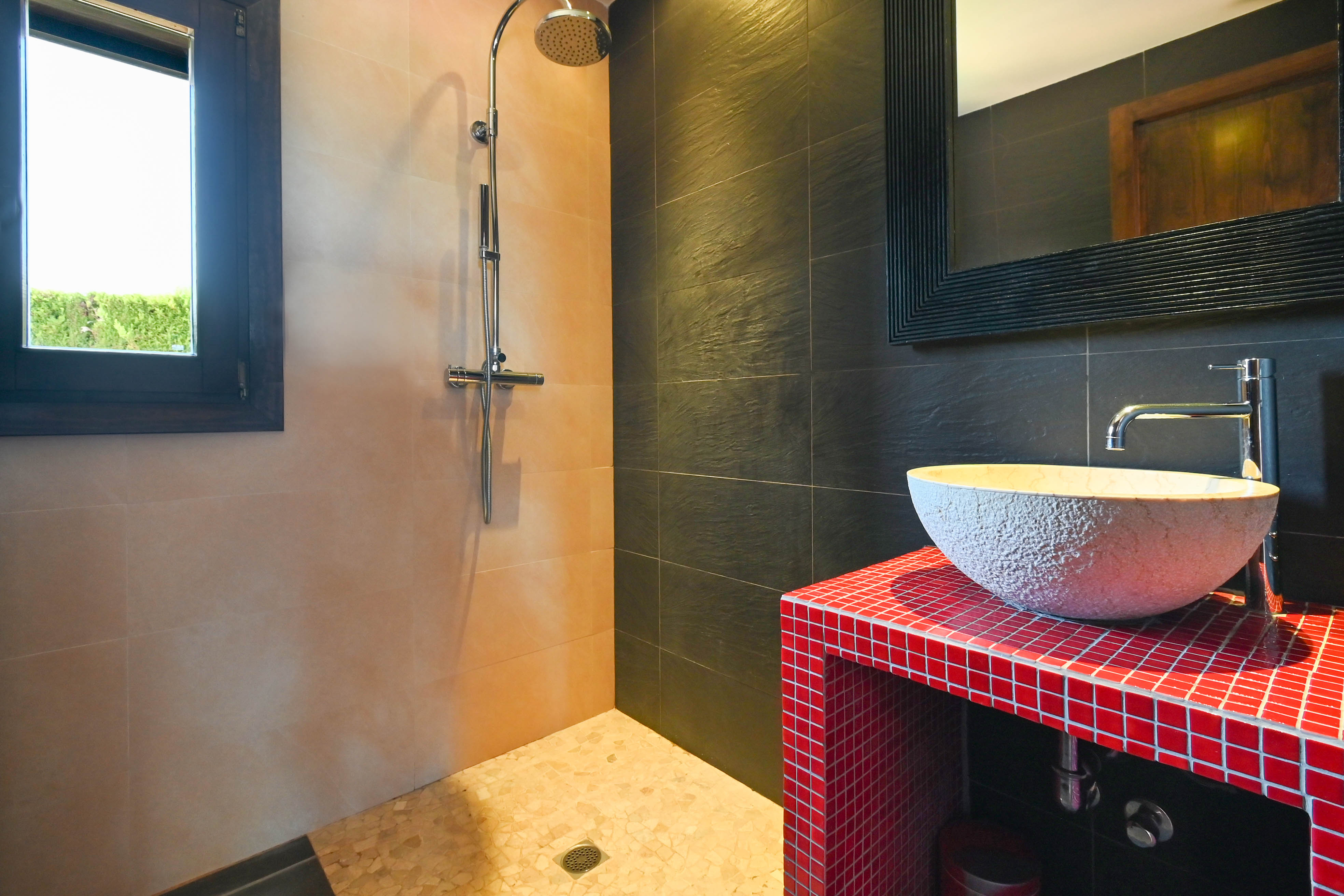 Badezimmerdetail mit Dusche, rustikalem Waschbecken auf roter Mosaikplatte und Fliesen in neutralen und dunklen Tönen.