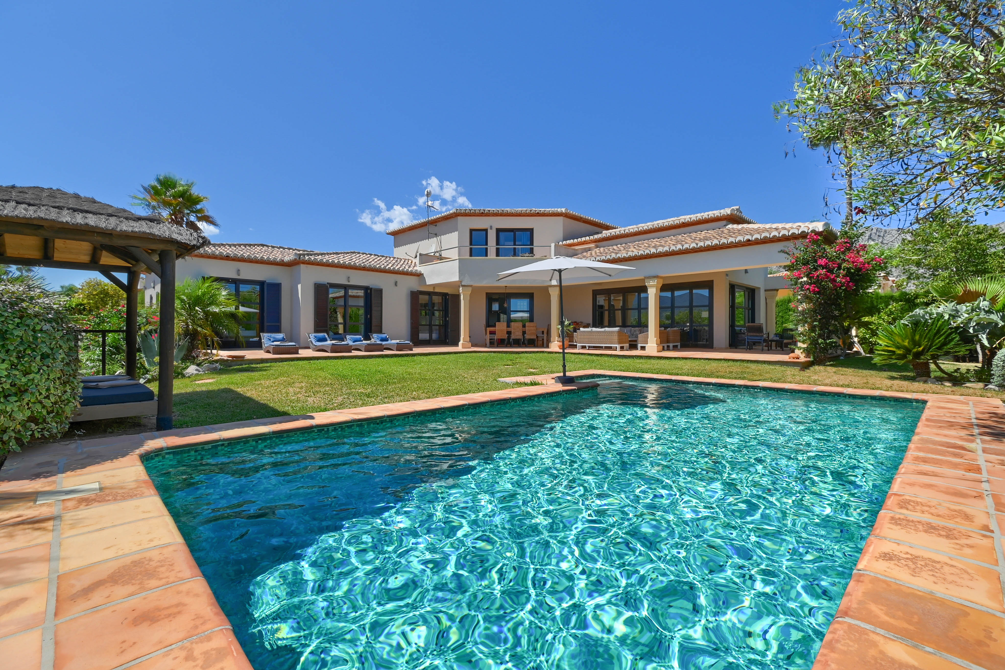 Blick auf den türkisfarbenen Pool mit Terrakotta-Einfassung und die zweistöckige Villa mit Fensterläden.