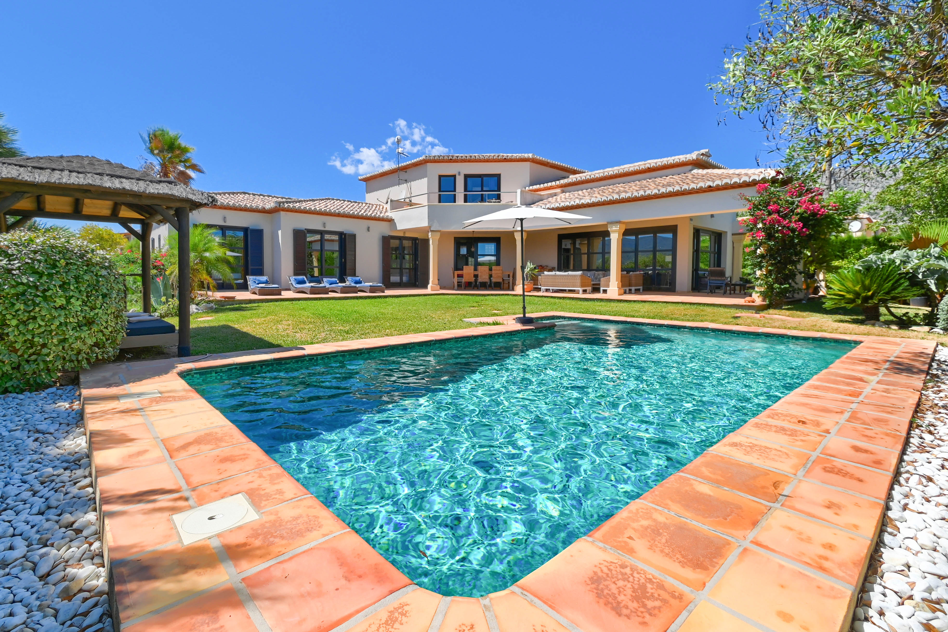 Blick auf den türkisfarbenen Pool mit Terrakotta-Rand und die Villa mit Garten und Pavillon.