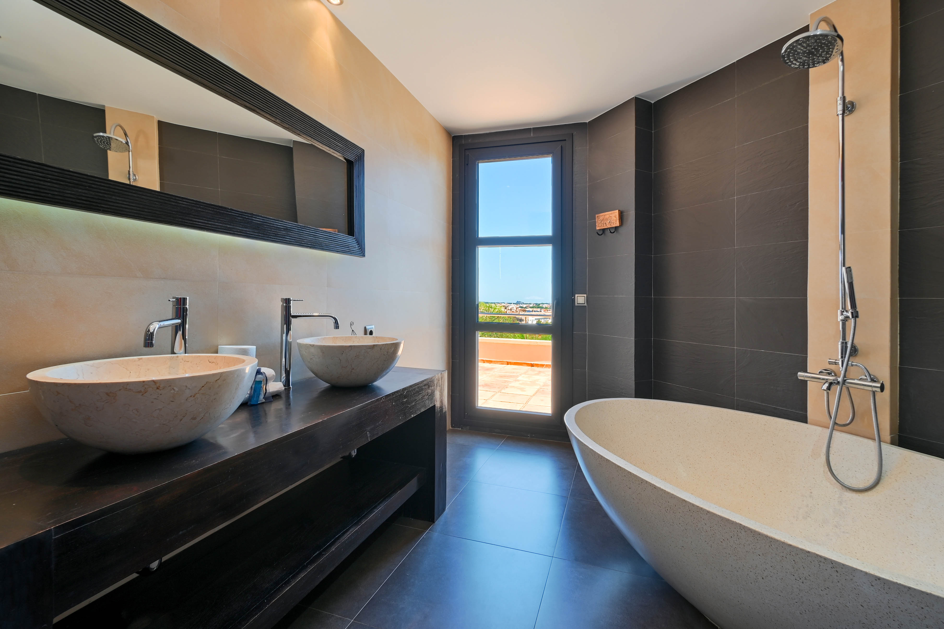 Elegantes Badezimmer mit Doppelwaschbecken aus Stein und freistehender Badewanne mit Zugang zu einer Außenterrasse.